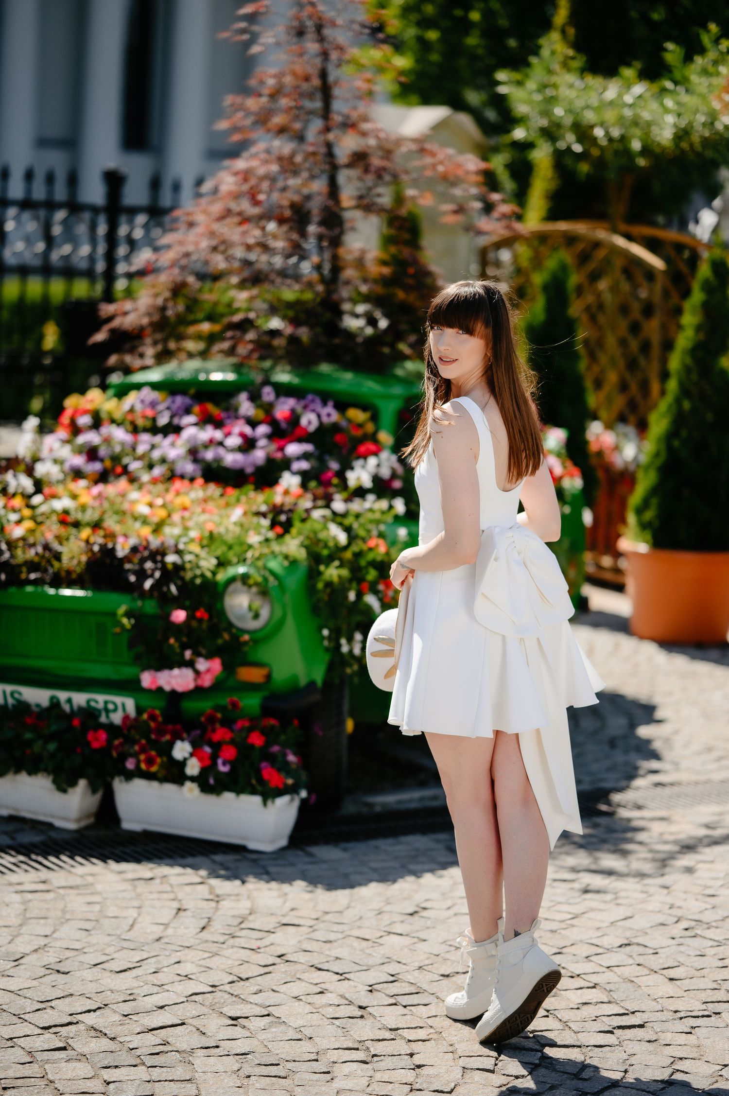 Young woman in white asymmetric dress posing beside a flower-decorated vintage green car on cobblestones.
