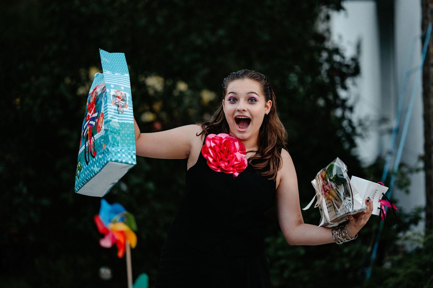 Excited young woman holding gift bags and flowers at an outdoor birthday celebration.