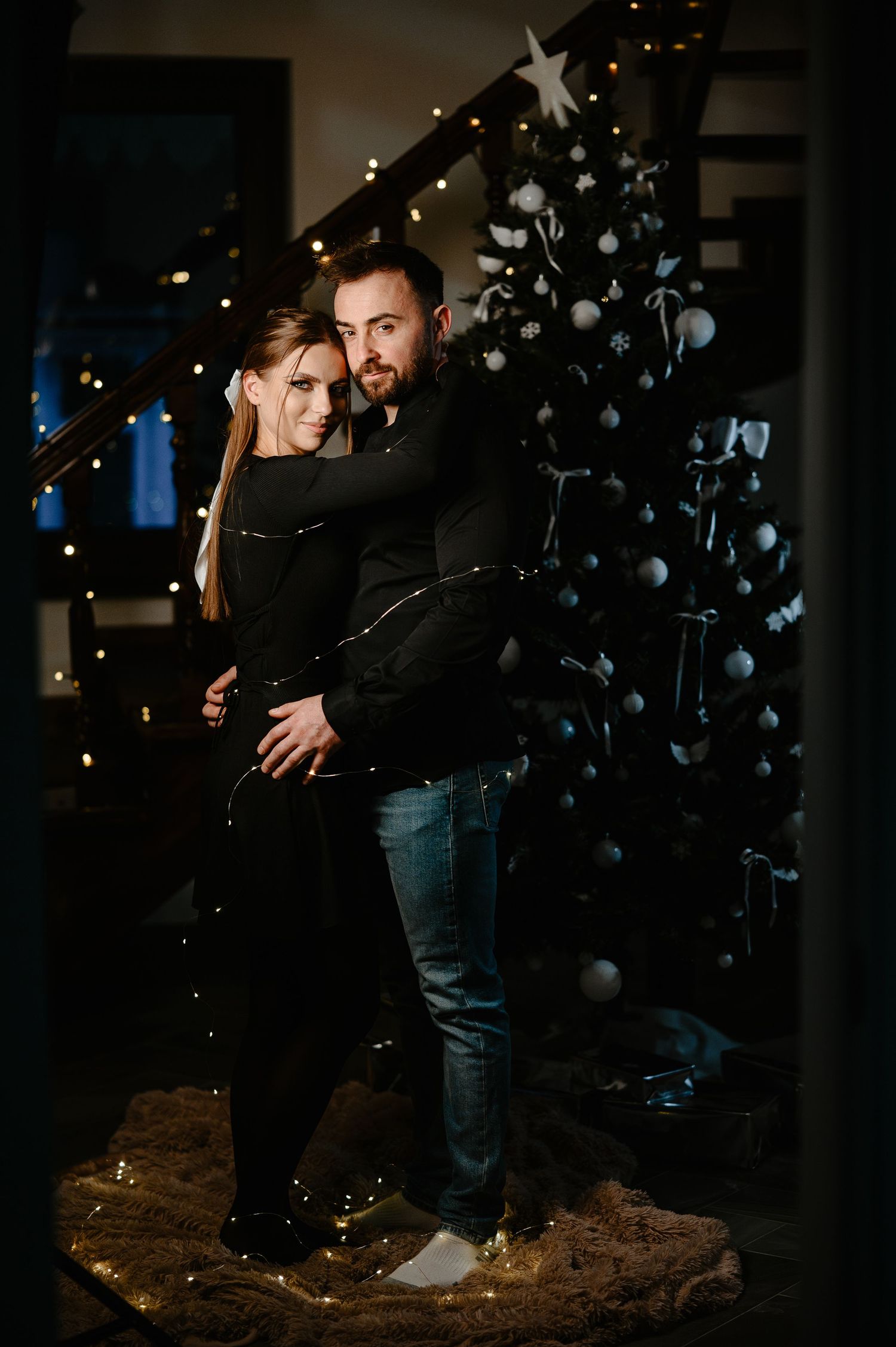 Couple hugging in front of a blue and white decorated Christmas tree in a dark room.