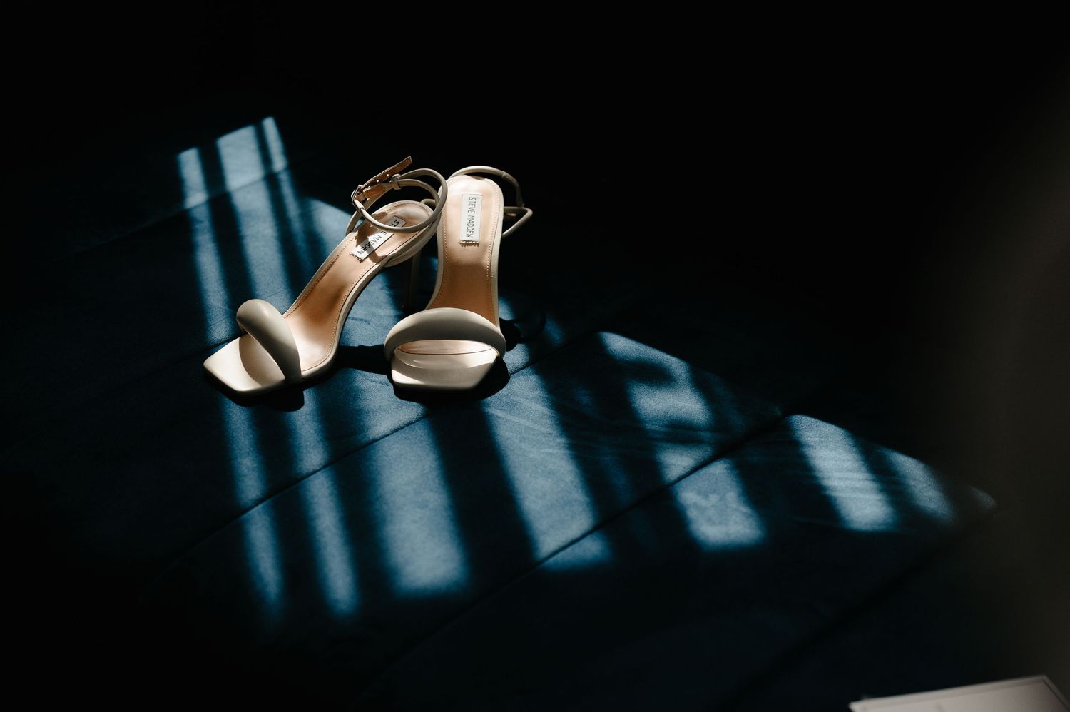 Gold strappy wedding heels casting dramatic shadows on dark floor in sunlight.