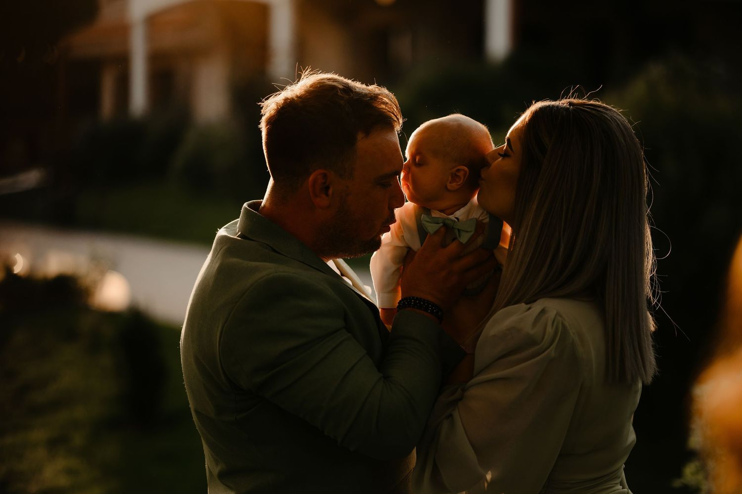 Parents in green attire sharing a kiss over their newborn baby in warm golden evening light.