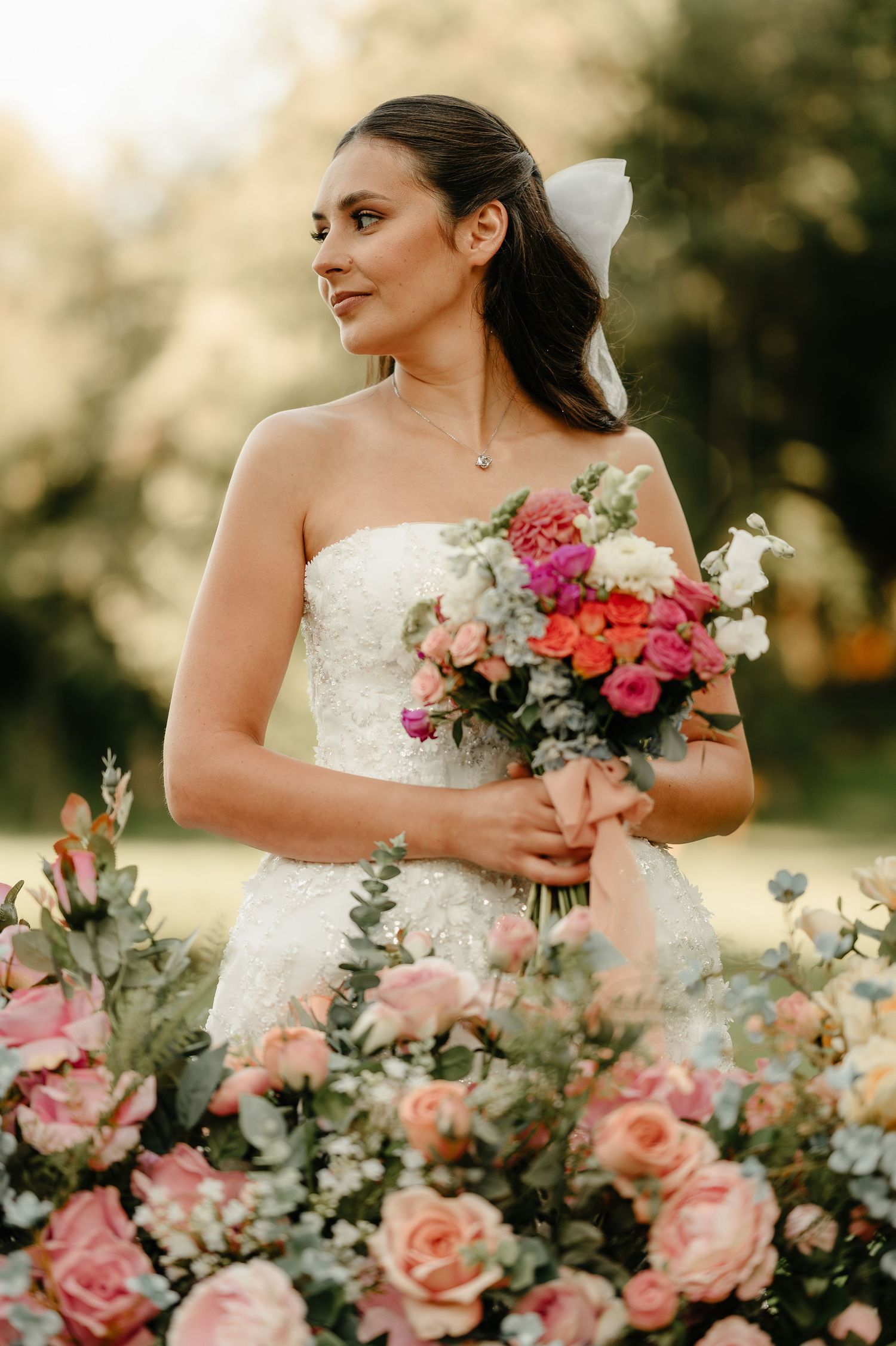 Elegant bride in strapless white gown holds vibrant bouquet surrounded by pink roses outdoors.