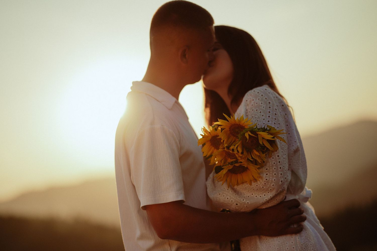 Couple sharing a kiss holding sunflowers together in a beautiful mountain sunset.