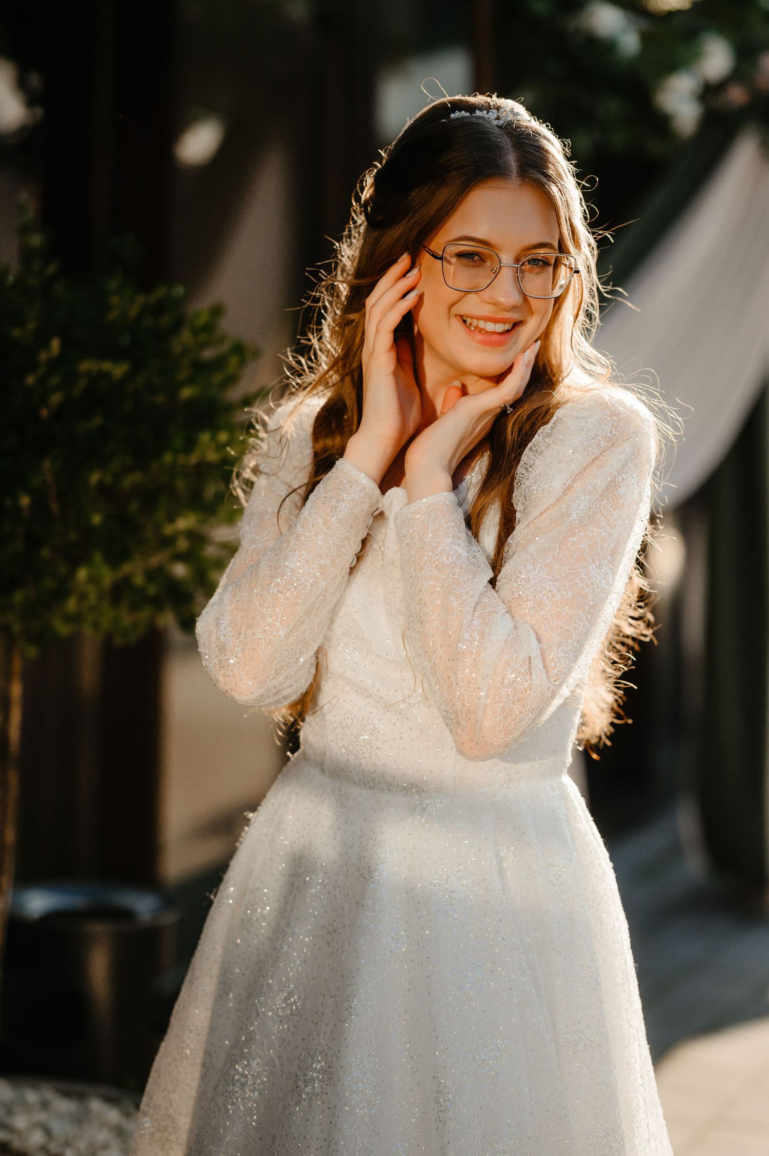 Radiant bride in glittery white wedding dress with glasses smiling in golden sunlight.