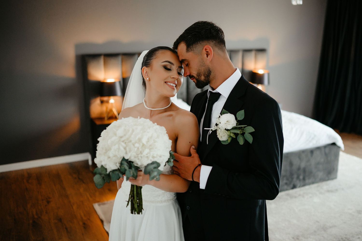 Bride and groom share an intimate moment, she holds white hydrangea bouquet in a warmly lit room.