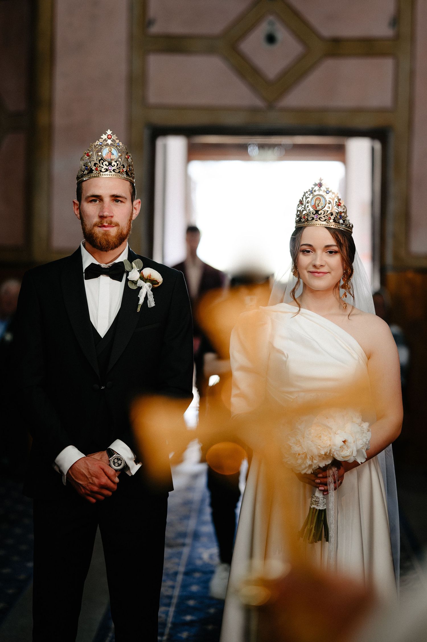Bride and groom wearing traditional Orthodox wedding crowns standing together in church aisle.