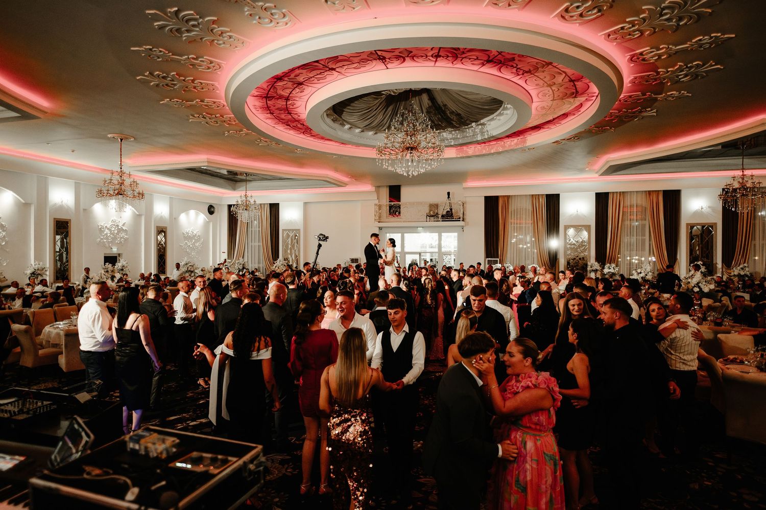 Elegant wedding reception ballroom filled with guests dancing under ornate ceiling with pink lighting.