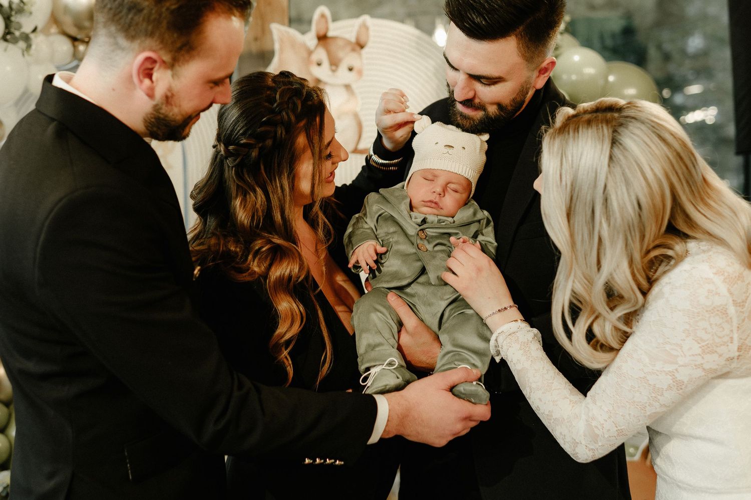 Two couples admiring a newborn baby dressed in sage green at an elegant woodland-themed christening celebration.