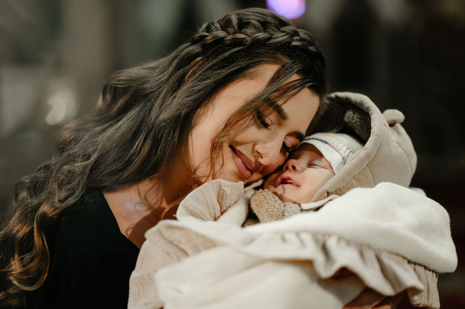 Mother with braided hair smiling lovingly at sleeping newborn in white baptism wrap.