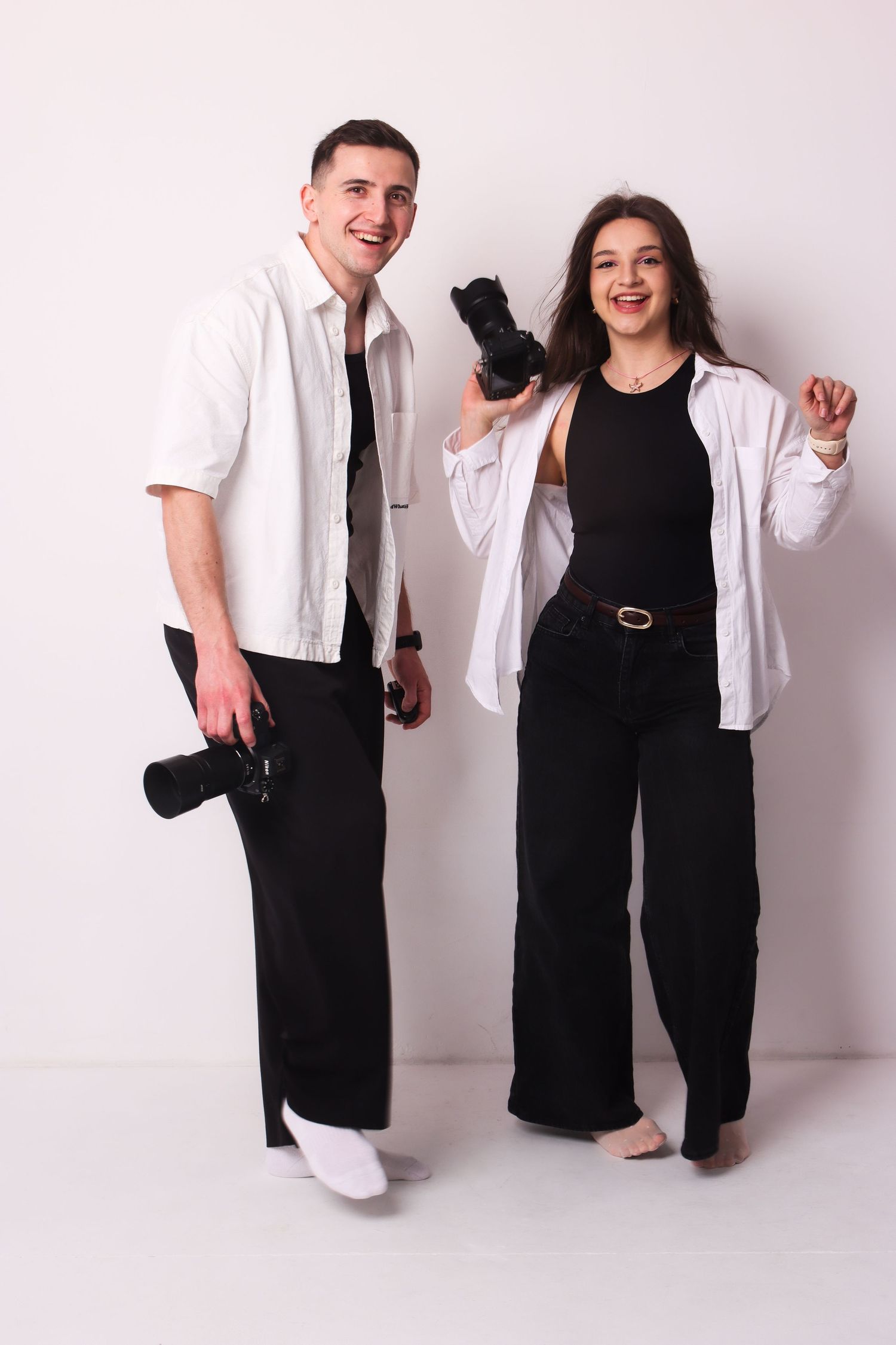 A smiling man and woman hold cameras while posing together against a white background.