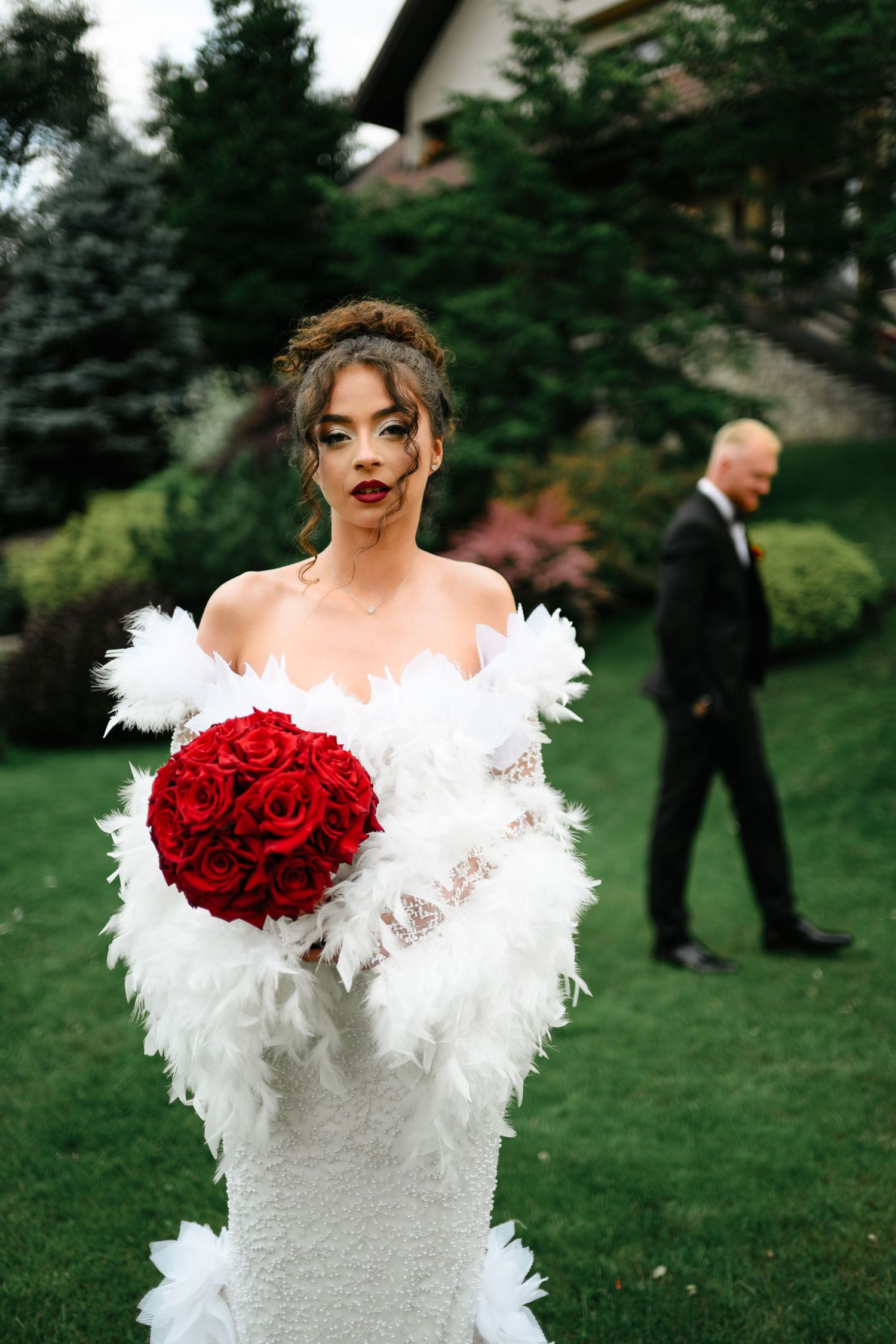 Bride in white feather wedding dress holds red rose bouquet outdoors, groom in black tuxedo stands in background.