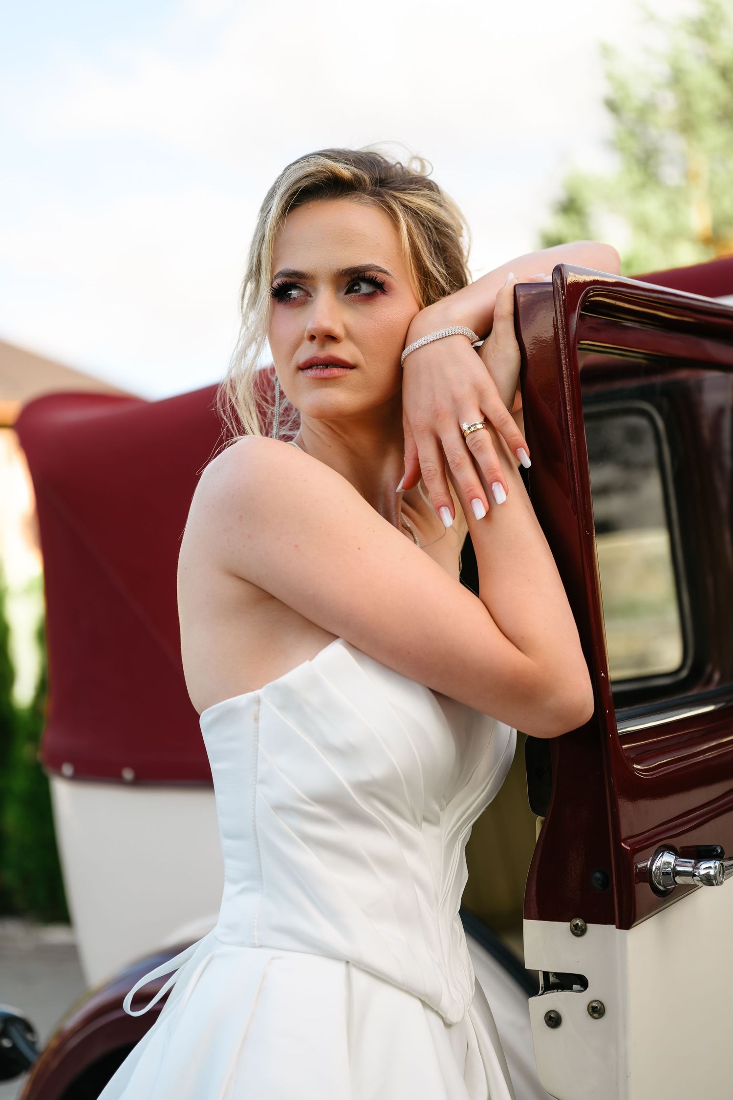 Elegant bride in white strapless gown leaning against vintage burgundy car, showcasing diamond ring, with soft outdoor lighting.