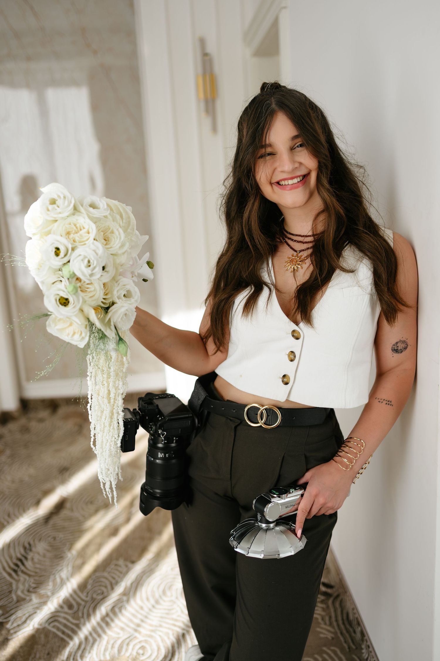 Smiling female photographer holding white flower bouquet and camera equipment, wearing white crop top and dark pants.