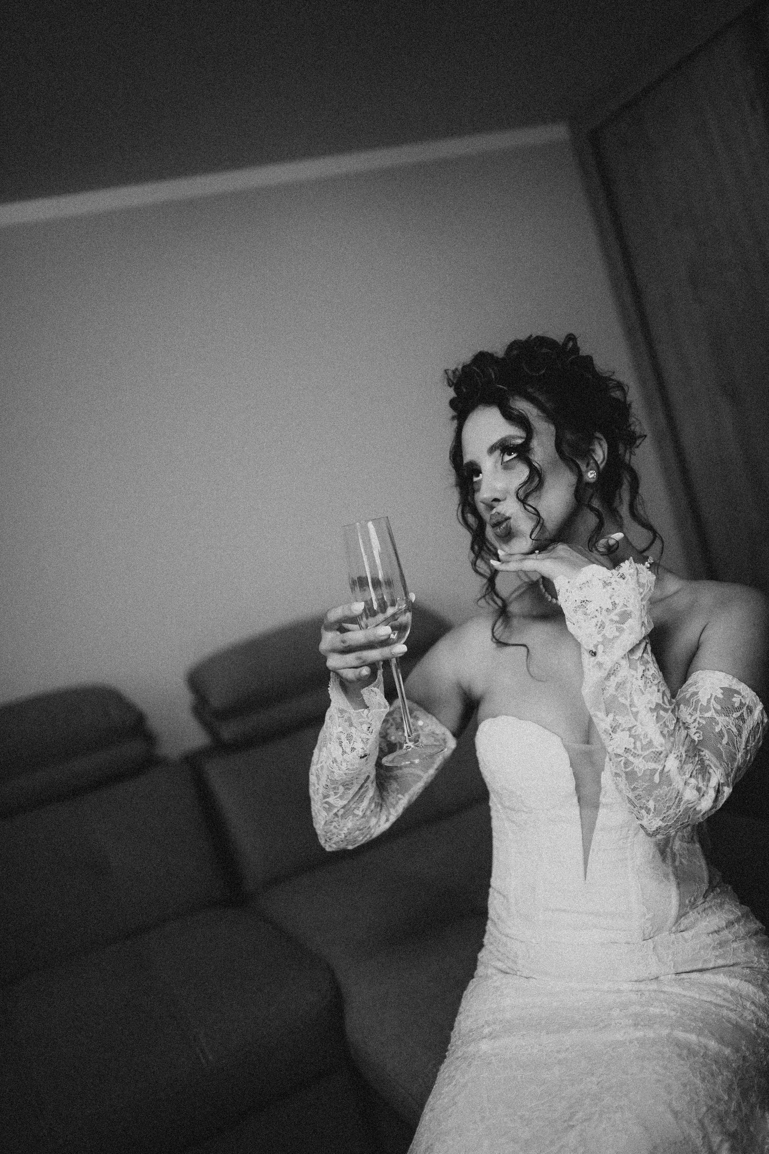 Black and white photo of a bride in lace wedding dress holding champagne glass, sitting near a sofa.