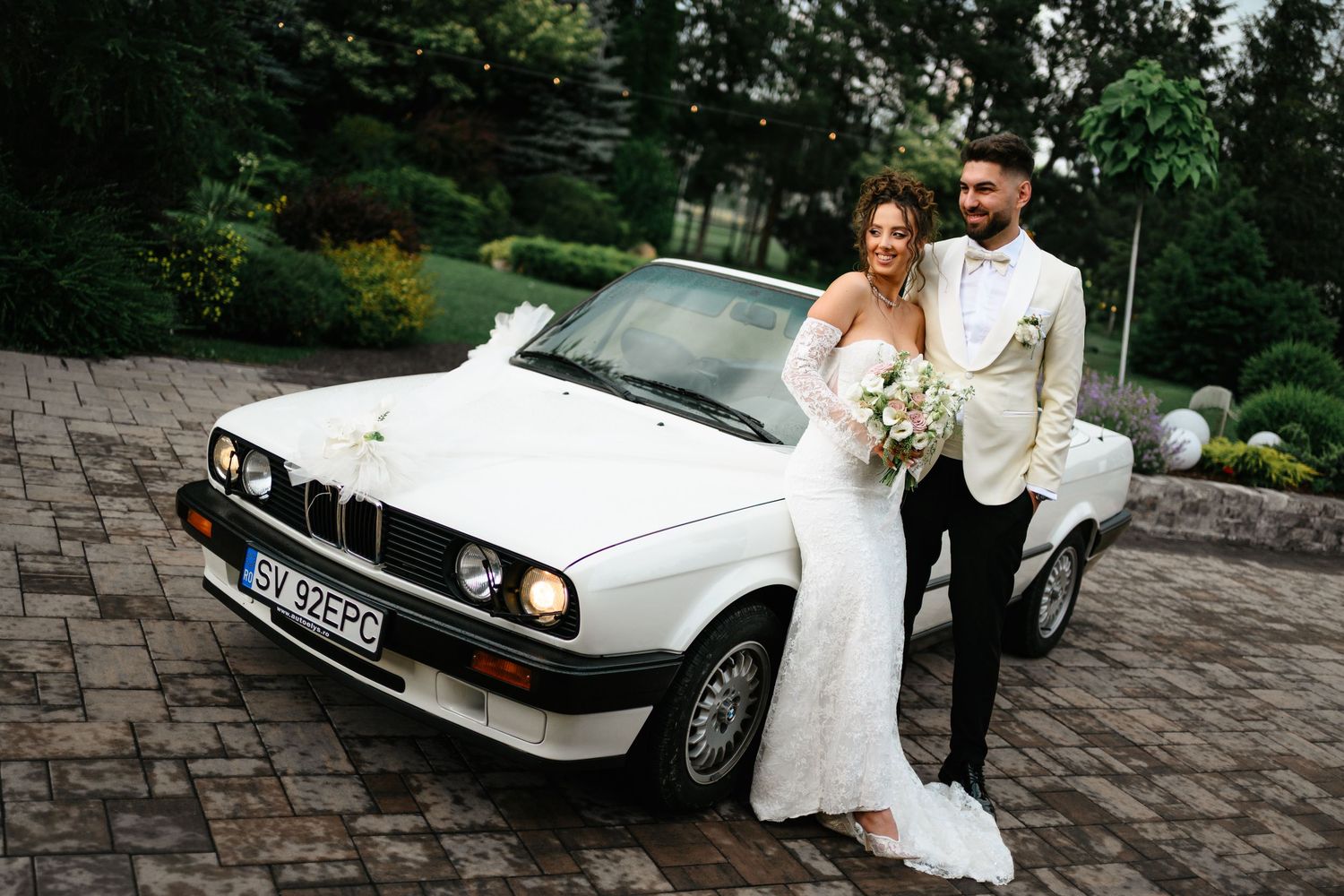 Bride and groom posing together in front of a classic white BMW convertible on their wedding day.