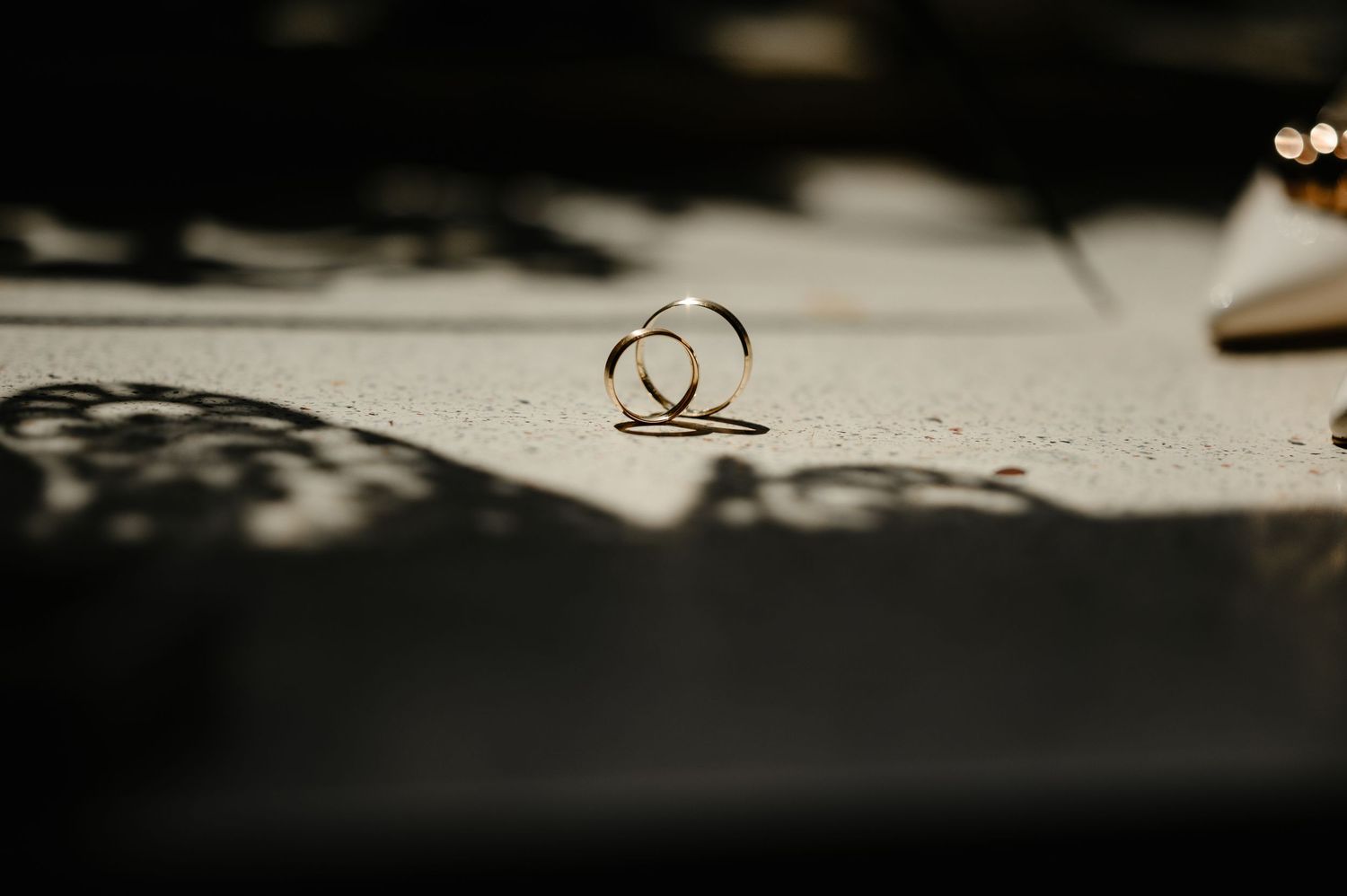 Two gold wedding rings standing upright on a white surface with dramatic shadows in soft bokeh light.
