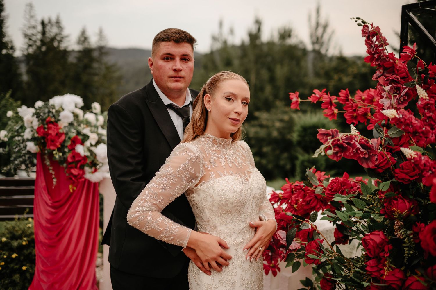 Wedding couple posing outdoors with red floral arch, bride in lace gown with groom in black tuxedo.
