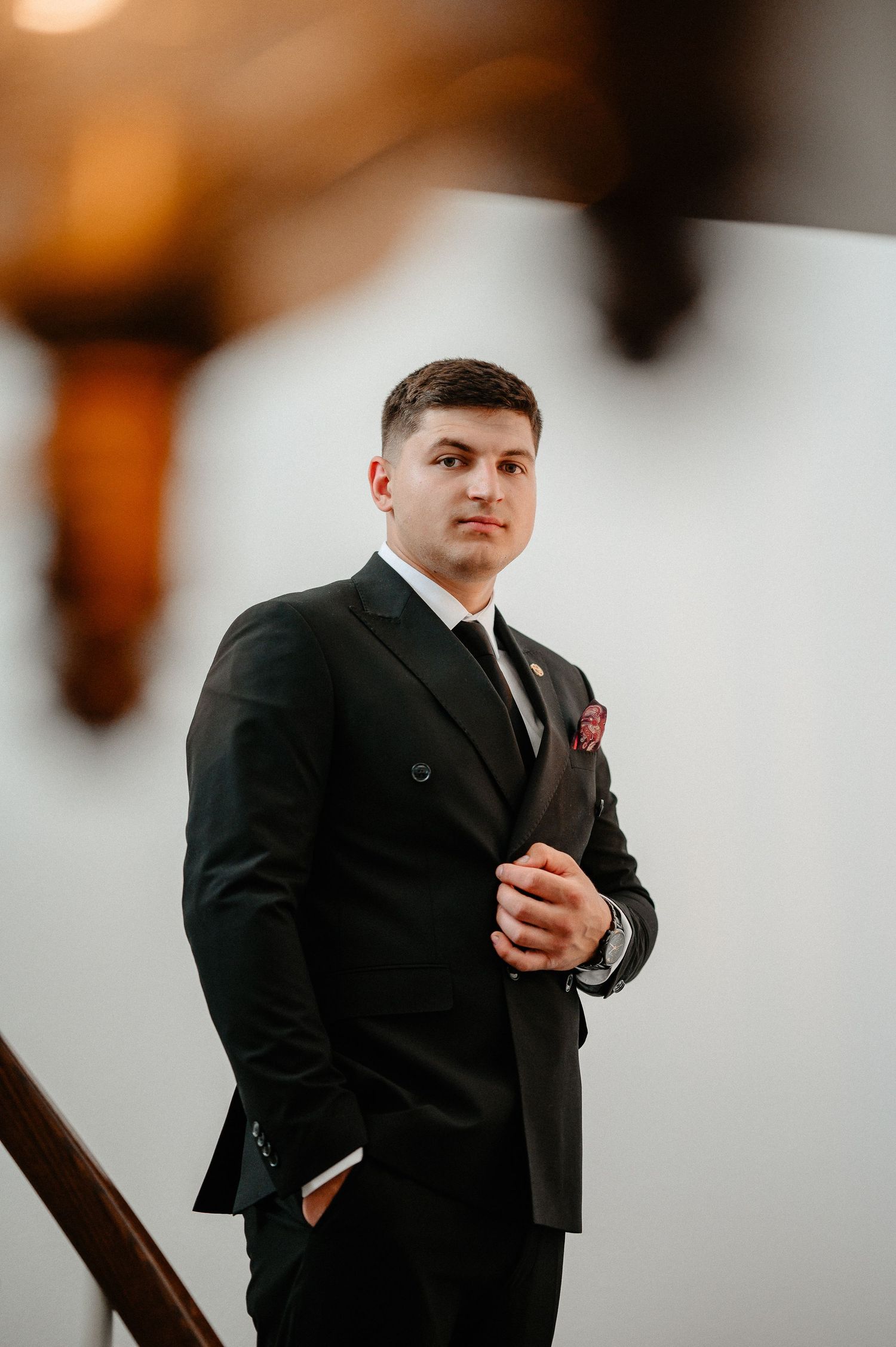 Groom in black double-breasted suit with red pocket square poses on staircase.
