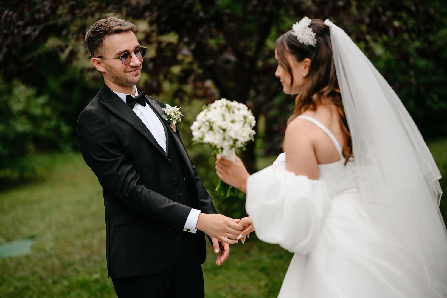 Groom in black tuxedo holds brides hands outdoors as she holds white wedding bouquet.