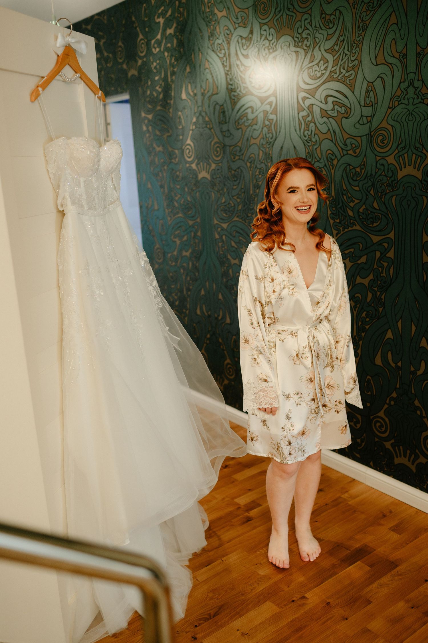 Smiling bride in floral robe stands barefoot beside her elegant white wedding gown hanging on a hanger against teal wallpaper.