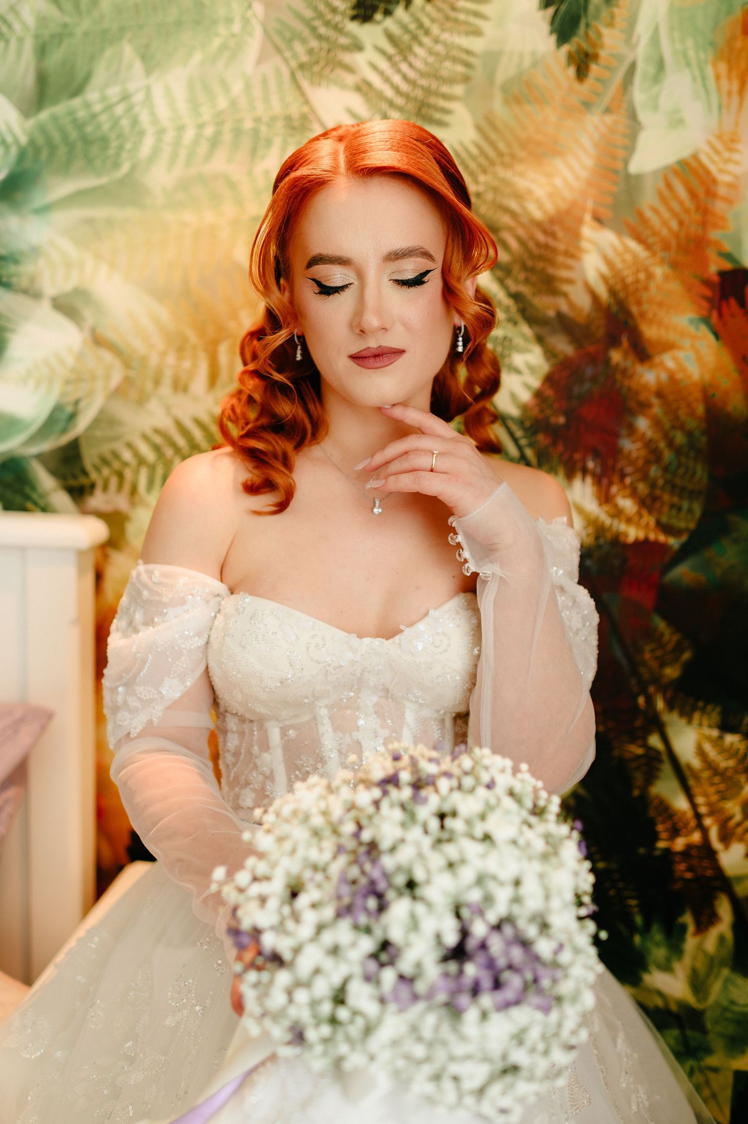 Red-haired bride in off-shoulder lace gown holds white floral bouquet before a tropical leaf backdrop.