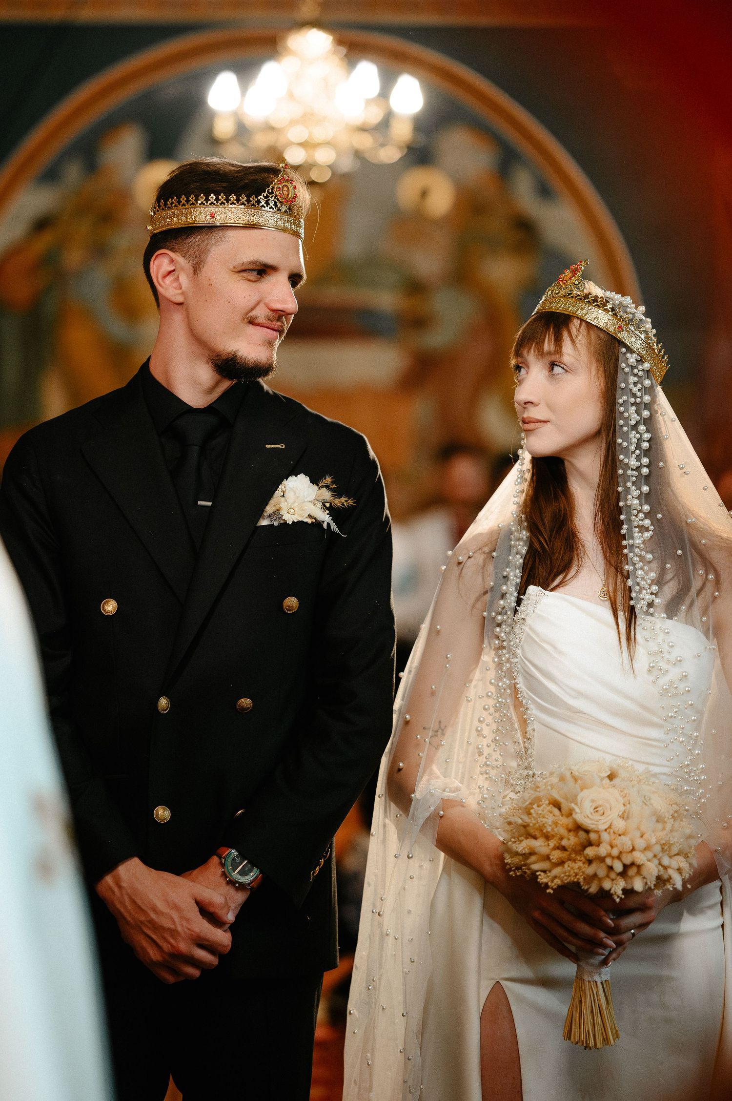 Bride and groom wearing ornate gold Orthodox wedding crowns stand together inside decorated church.