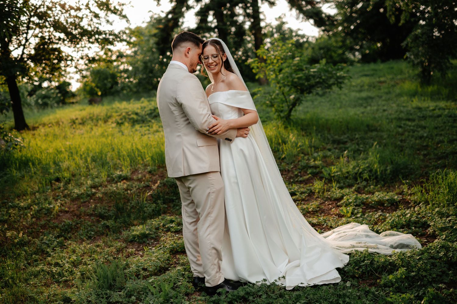 Bride and groom embracing in lush green meadow at golden hour during wedding portraits.