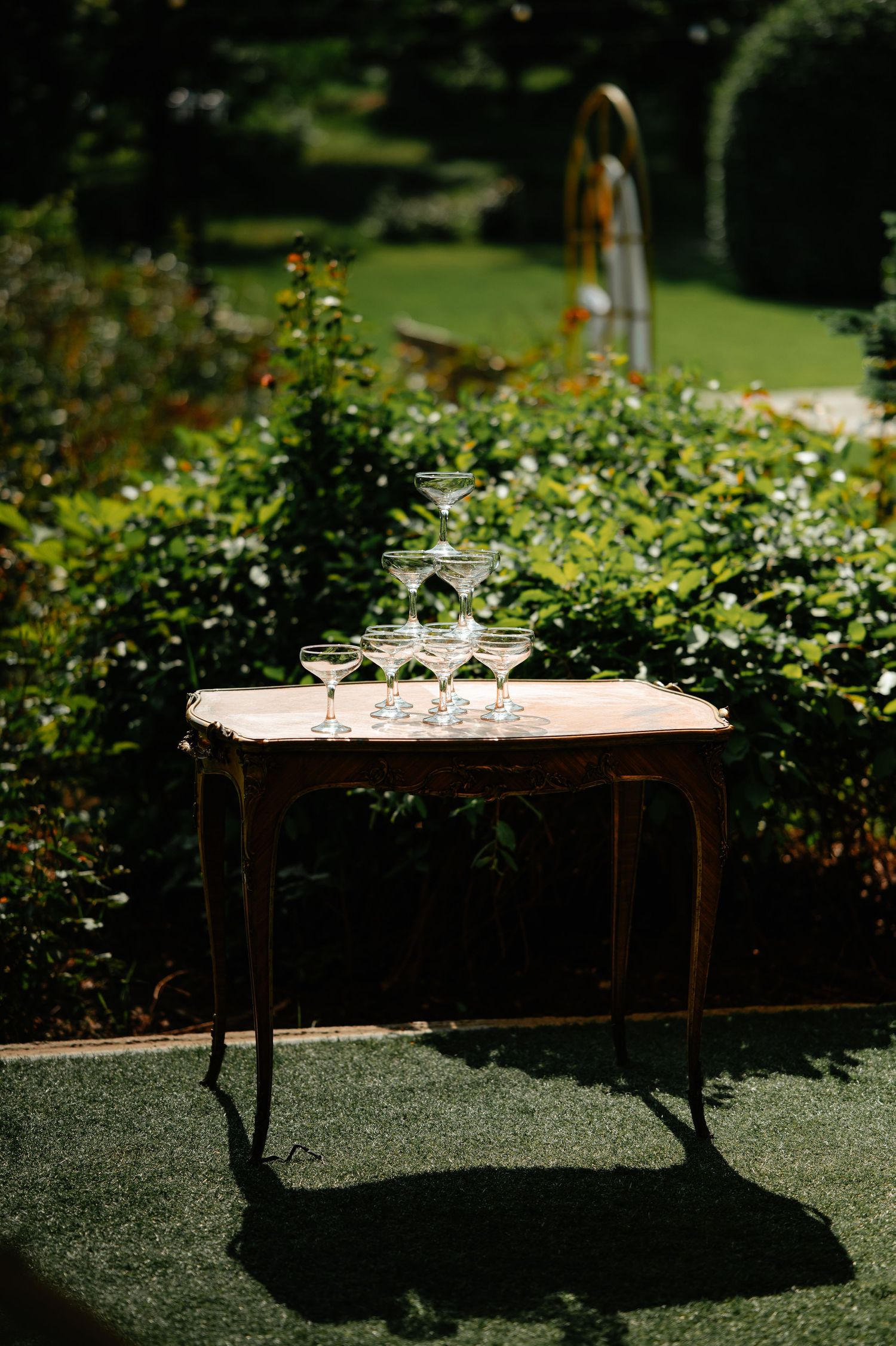 Champagne coupe glasses stacked in pyramid on antique wooden table in sunlit garden wedding setting.