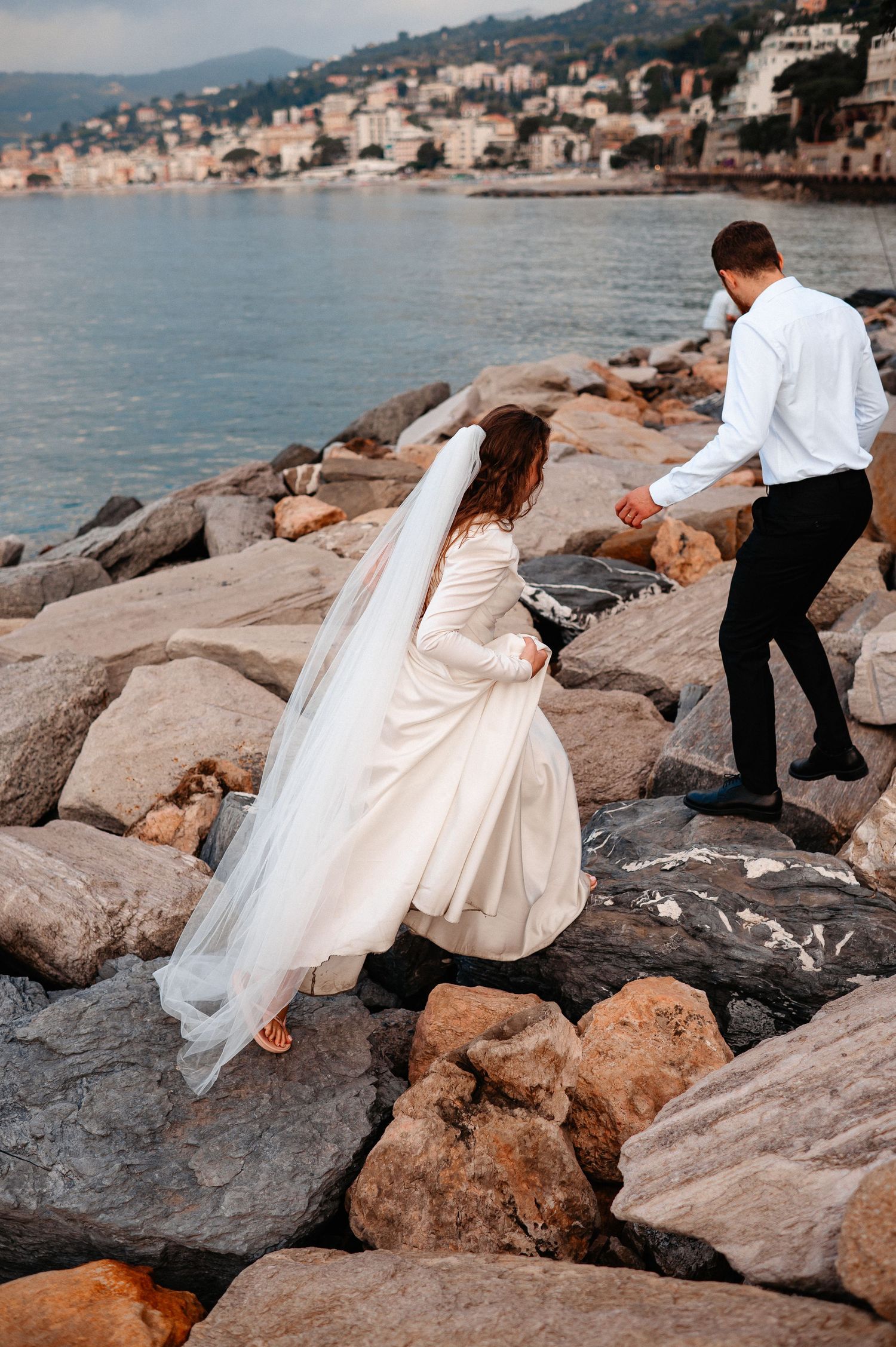 Bride and groom walk hand in hand over rocky shoreline with coastal town visible across the water.