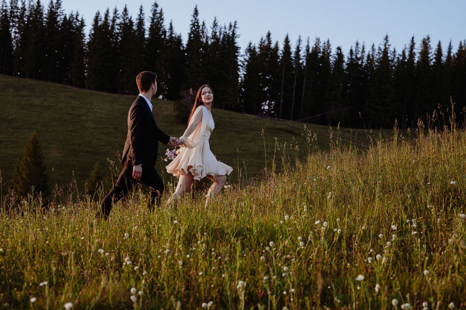 Bride and groom walking hand in hand through a sunlit mountain meadow surrounded by evergreen trees.