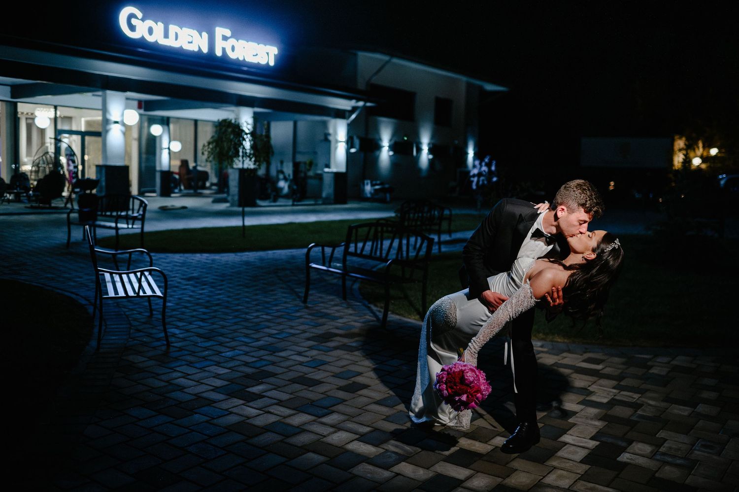 Groom dramatically dipping bride outside Golden Forest wedding venue at night with pink bouquet.