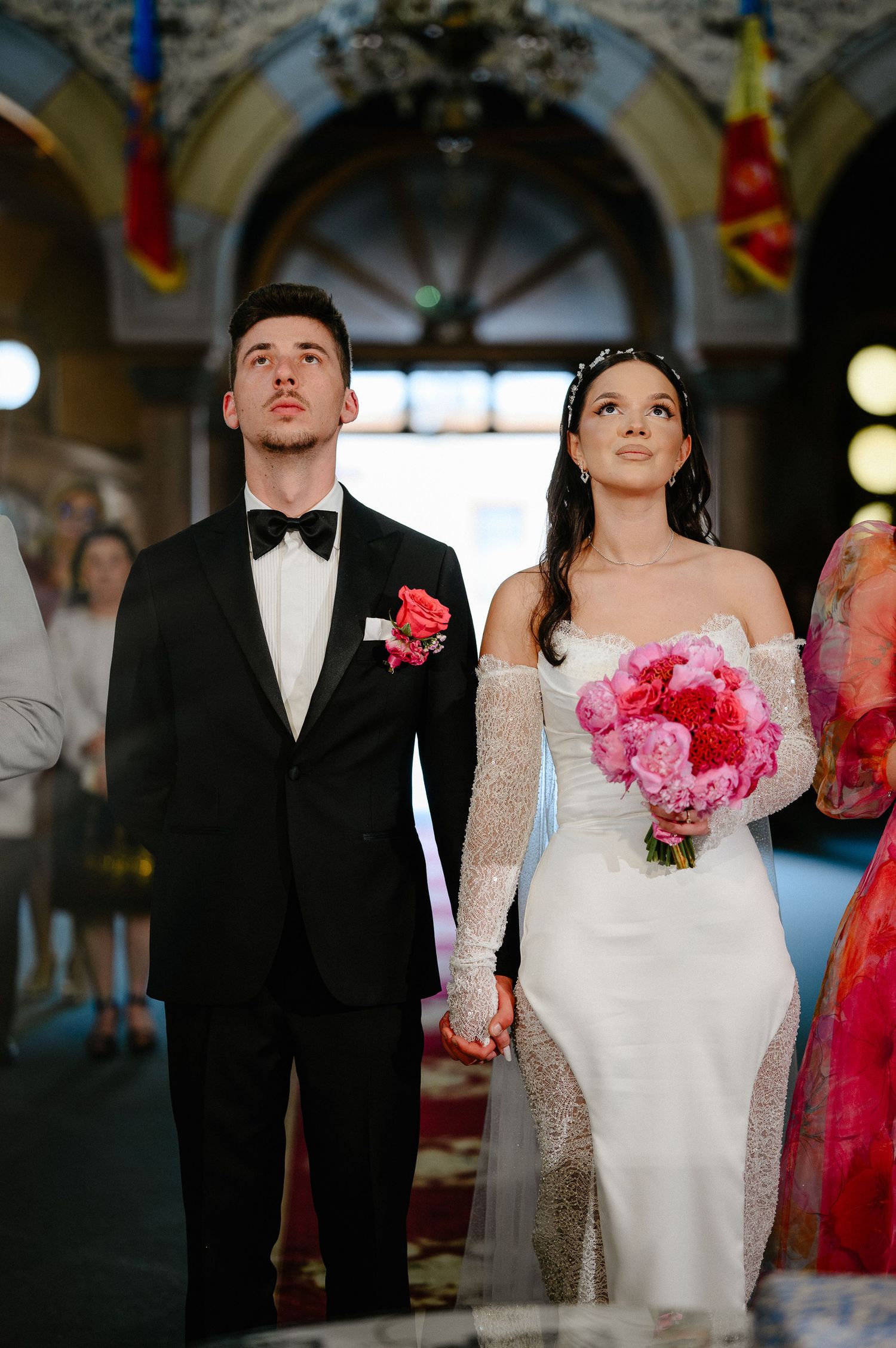 Bride and groom hold hands at Orthodox church altar, bride gazing upward with pink peony bouquet.