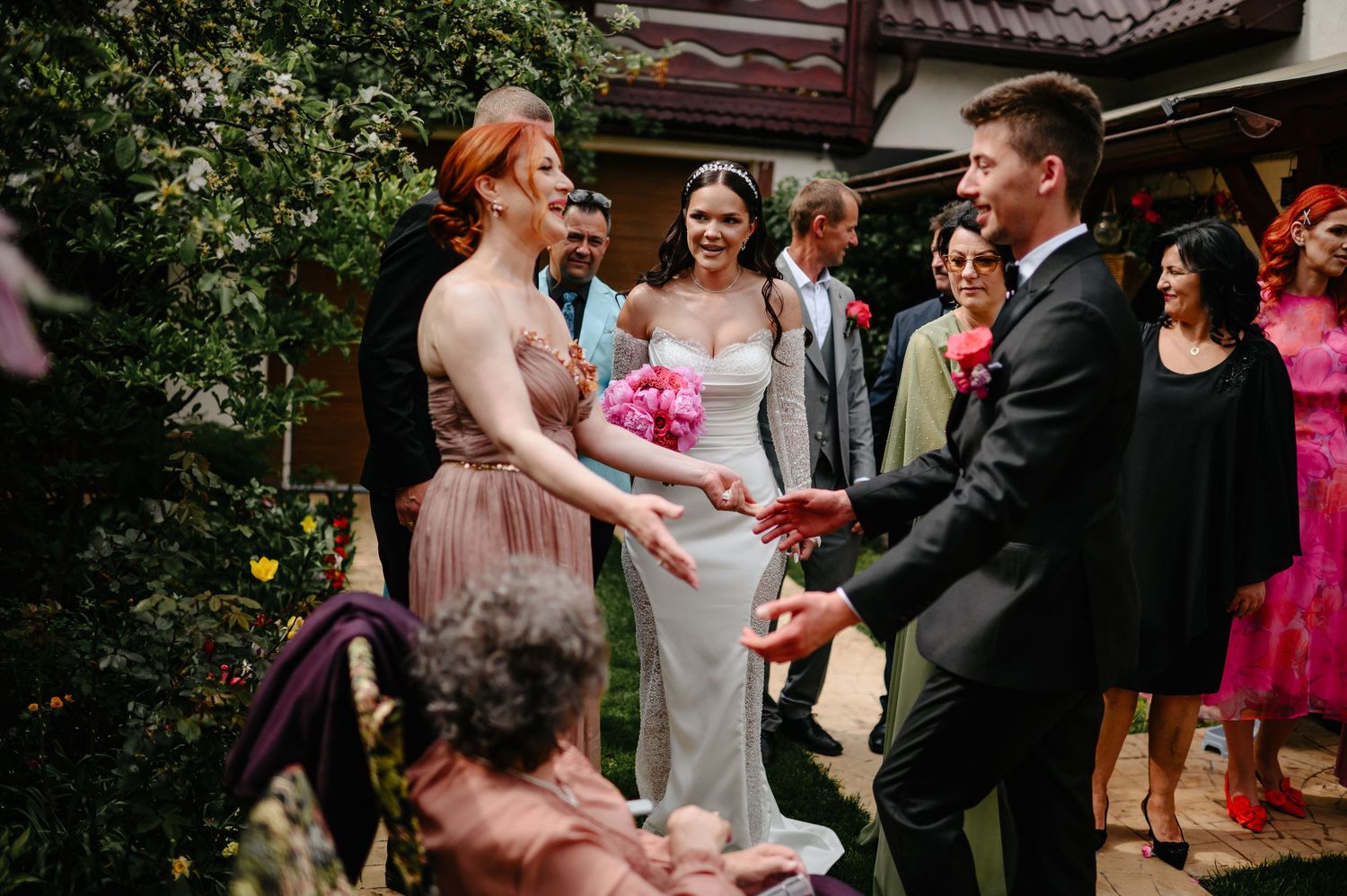 Bride in white gown with pink bouquet greeted by guests including redhead bridesmaid at outdoor wedding.