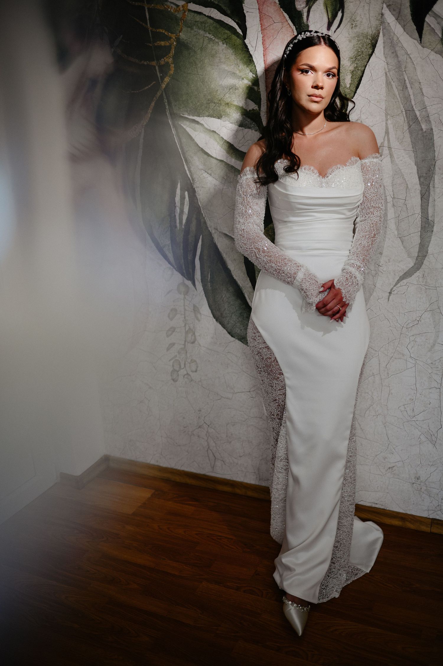 Elegant bride in off-shoulder lace wedding gown with beaded headpiece standing before tropical floral wallpaper.