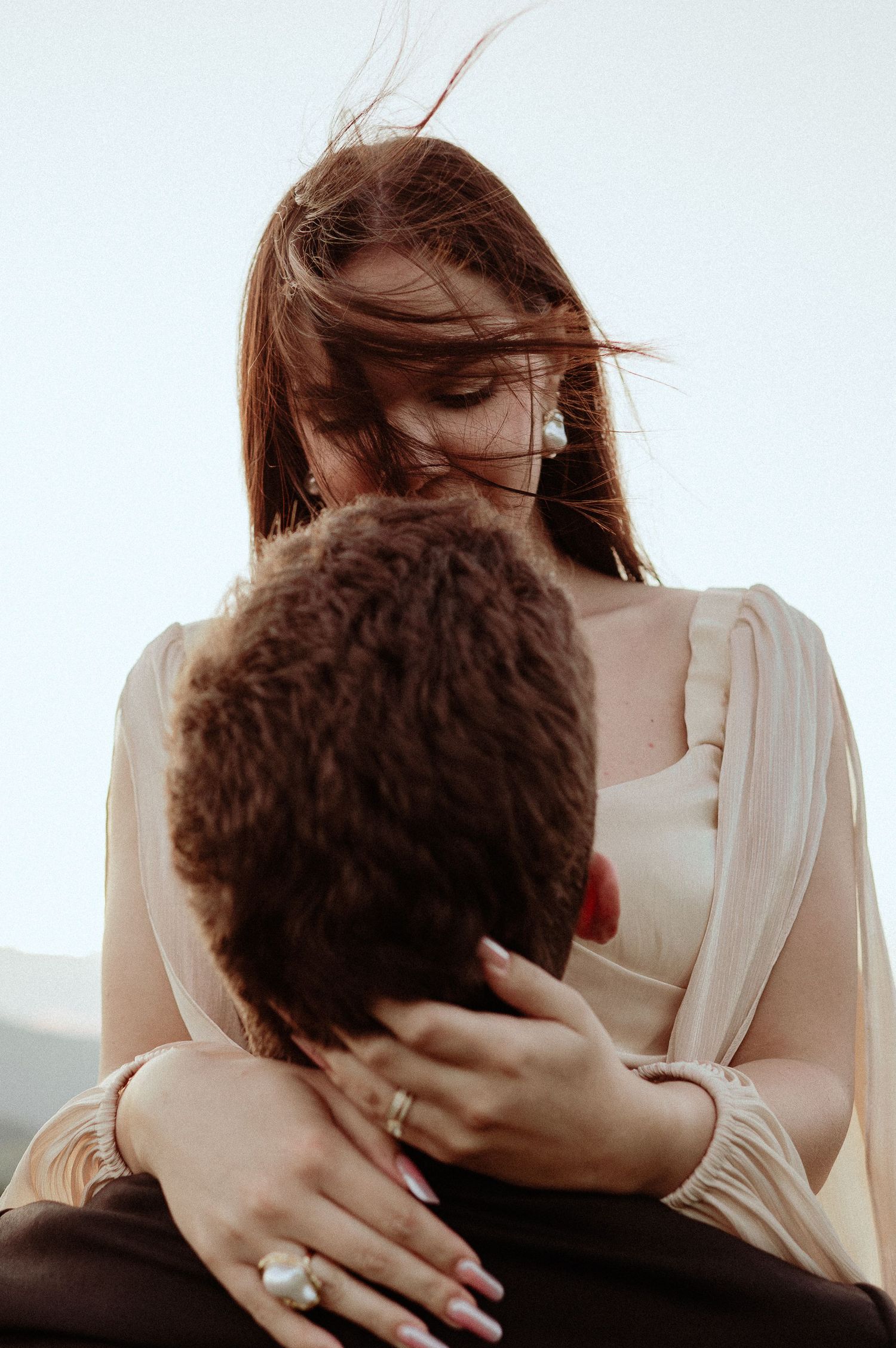 A woman with windswept red hair tenderly embraces a man while wearing a pearl earring and ring.