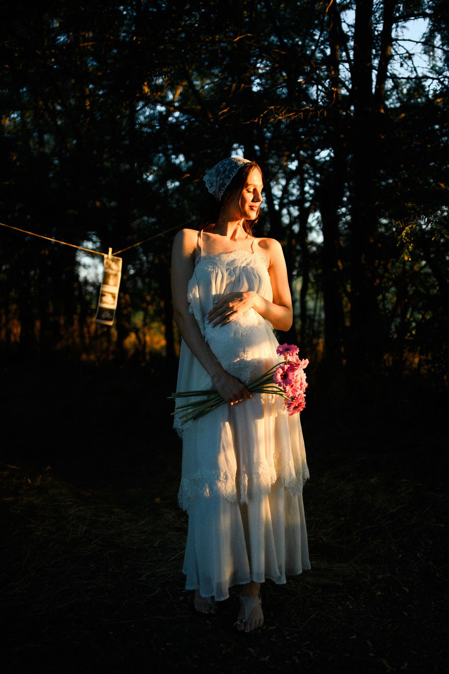 Pregnant woman in white dress holding pink flowers, bathed in golden sunset light in a forest setting.
