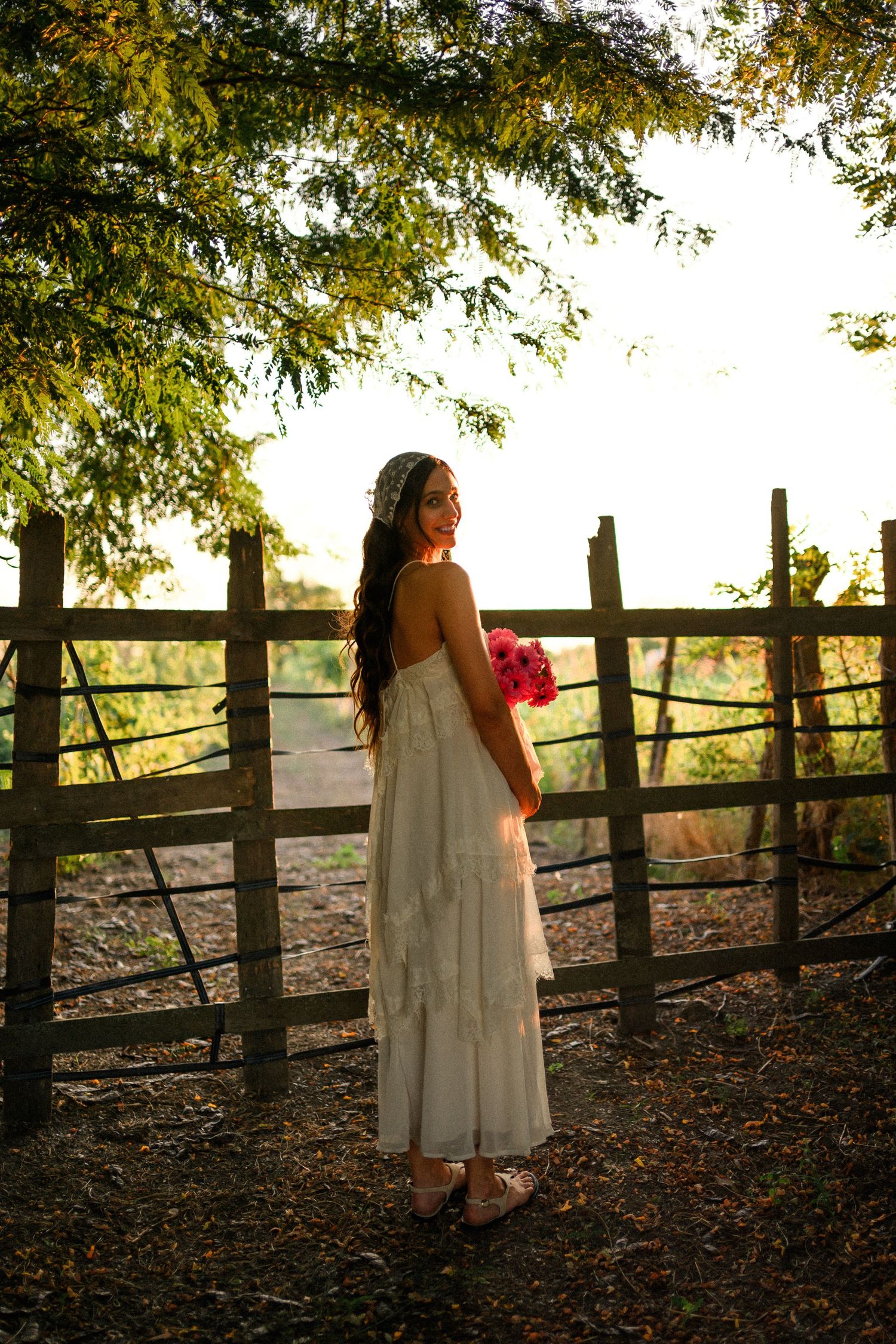 Bride in white dress holding pink bouquet stands by rustic wooden fence at golden hour.