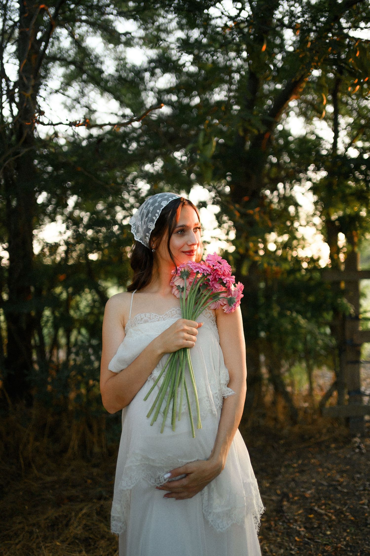 Pregnant woman in white dress holding pink flowers in a forest at golden hour.