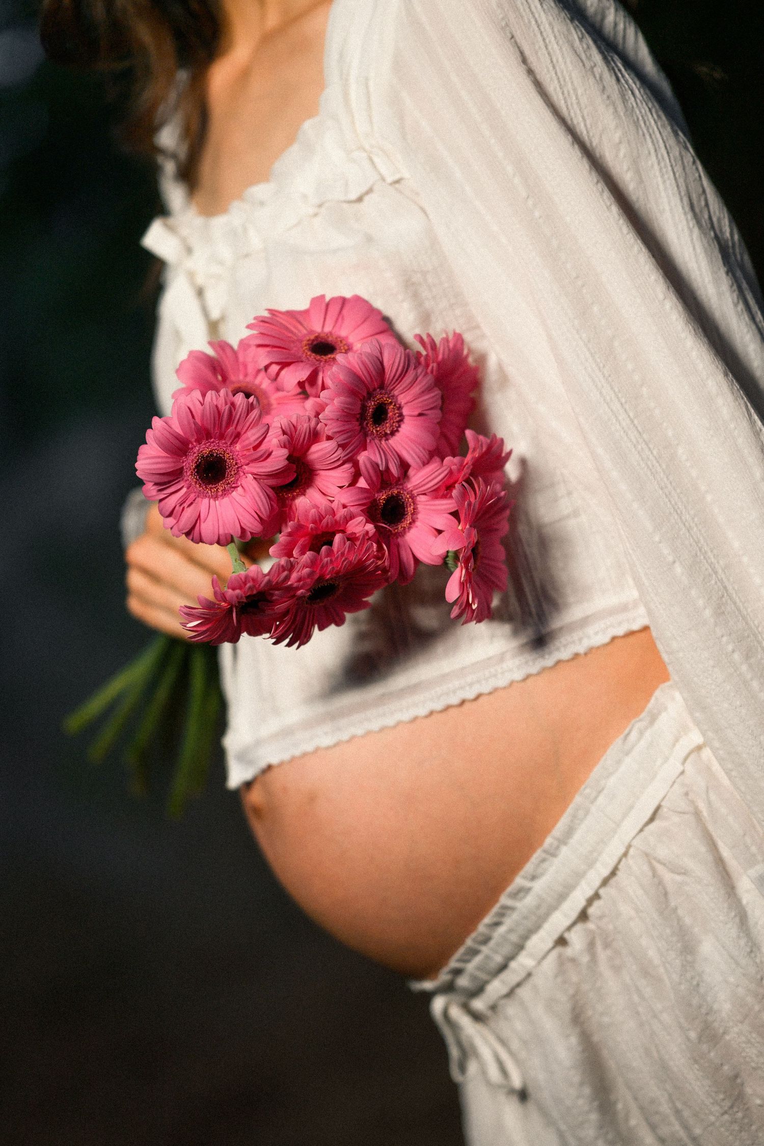 Pregnant woman in white dress holding a bouquet of pink gerbera daisies, showcasing her baby bump outdoors.