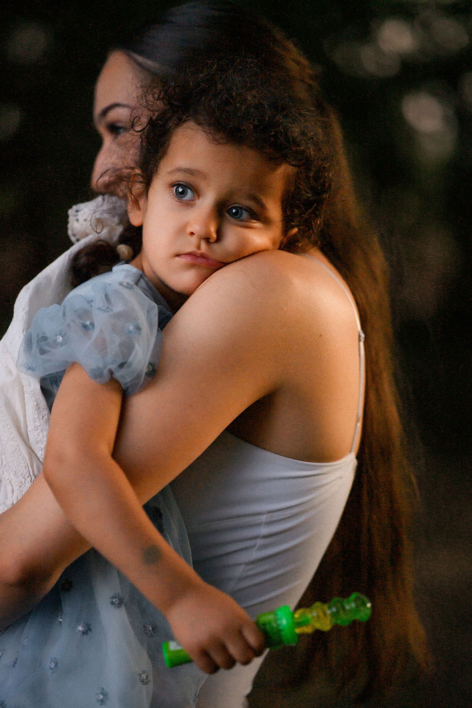A young girl with curly hair rests her head on a woman's shoulder while holding a green toy outdoors.