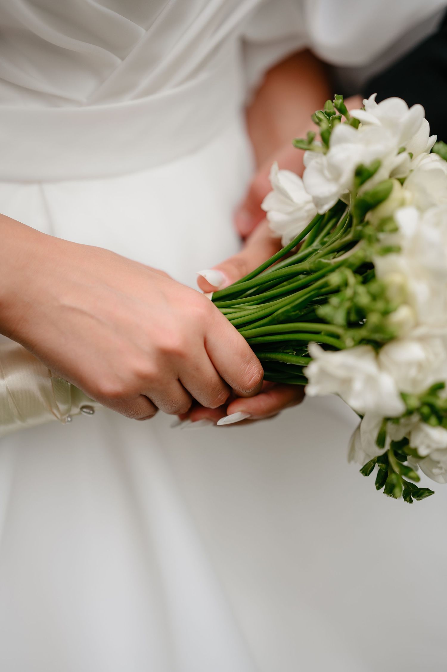 Bride holding elegant white freesia wedding bouquet with green stems against white wedding dress.