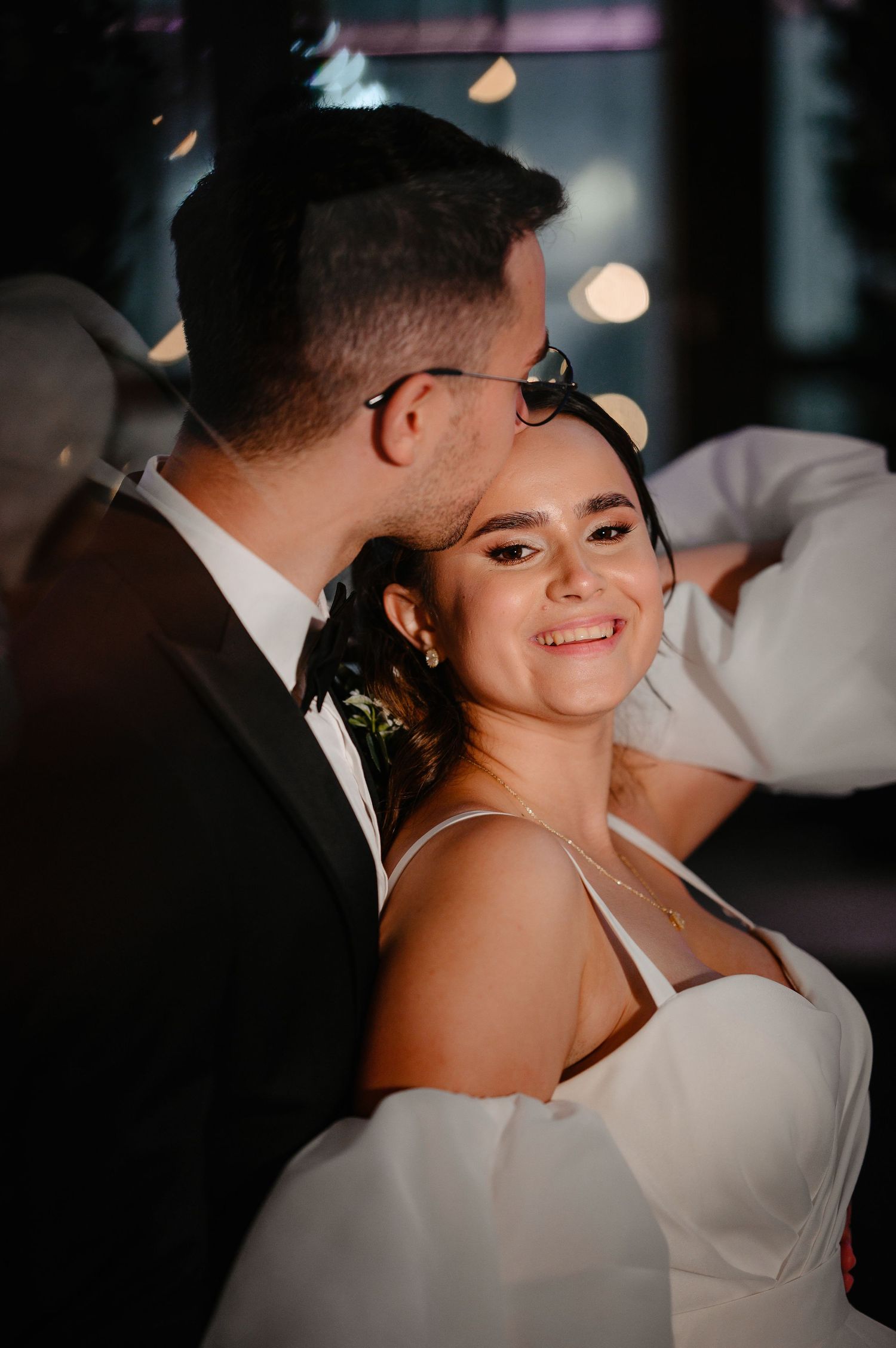 Groom kisses brides cheek as she smiles warmly at wedding reception with bokeh lights in background.
