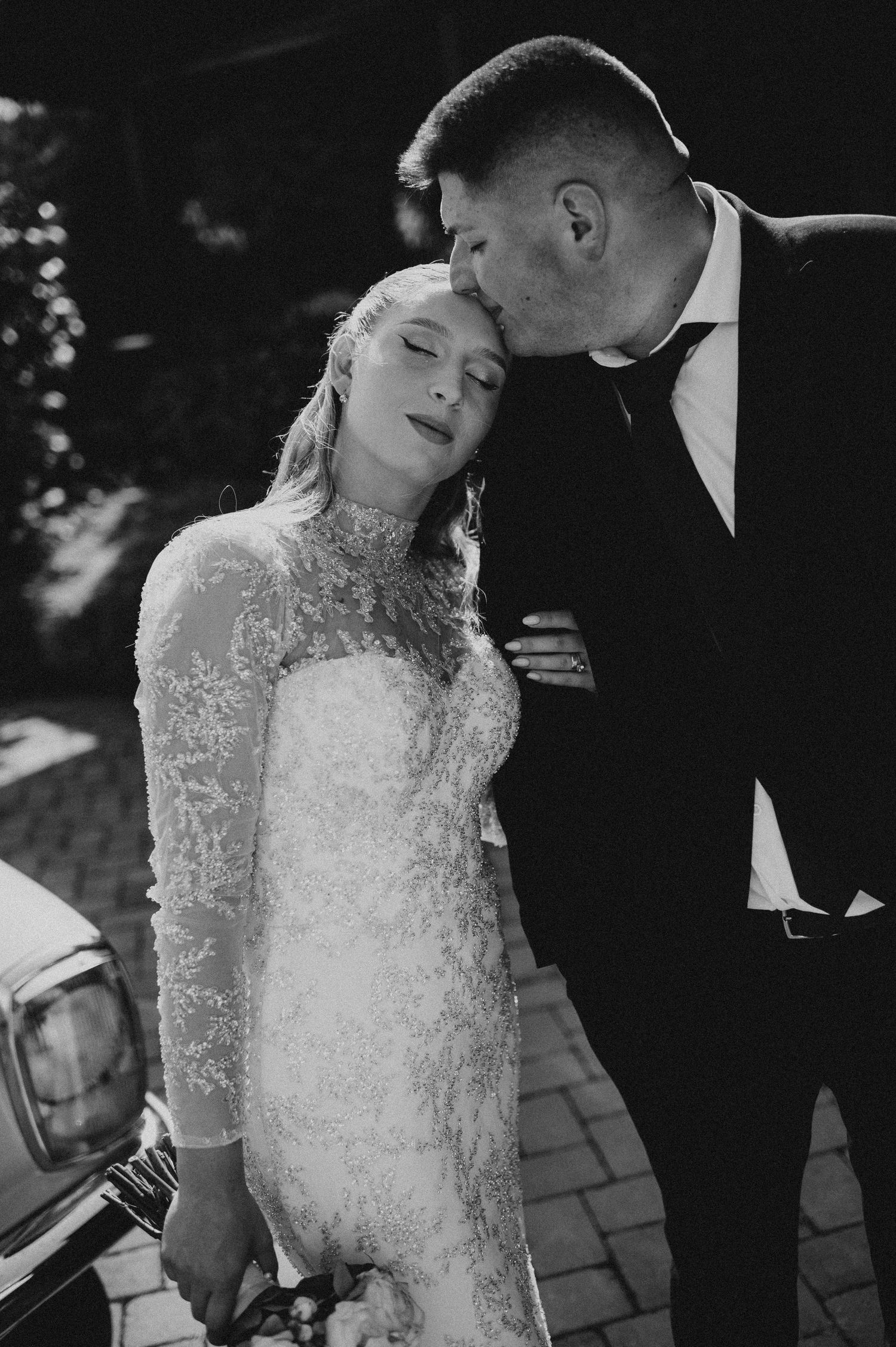 Black and white wedding photo of bride in lace gown and groom in tuxedo sharing a tender moment.