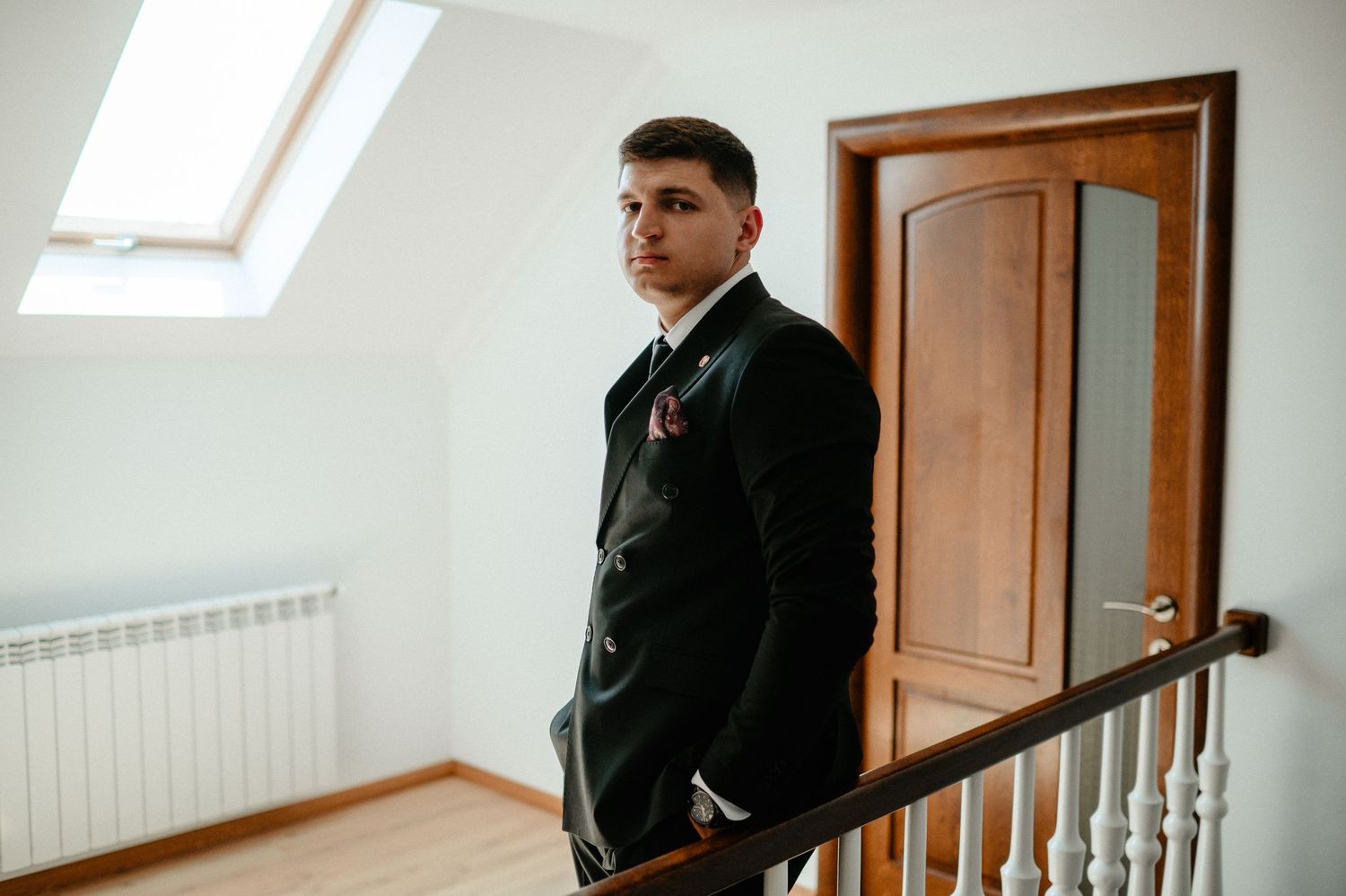 Groom in black suit standing on staircase near wooden door in bright white room before wedding ceremony.