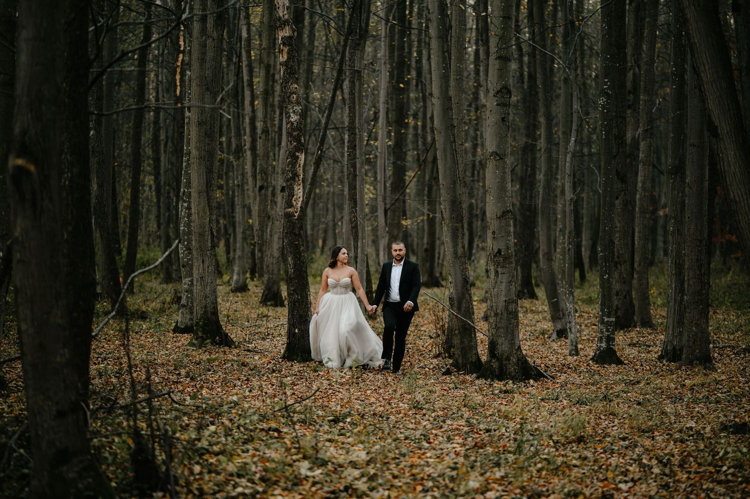 Bride and groom holding hands walking through a moody autumn forest with tall trees and fallen leaves.