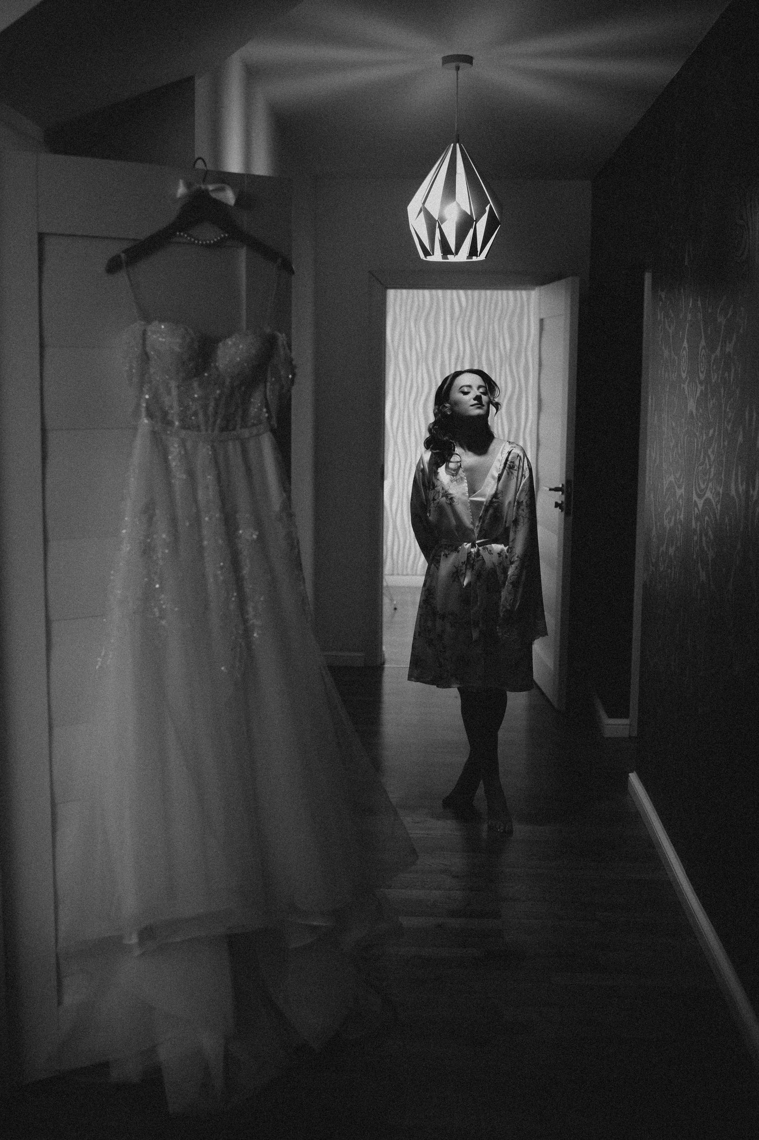 Bride in robe gazing at her wedding gown hanging in hallway, black and white photo.