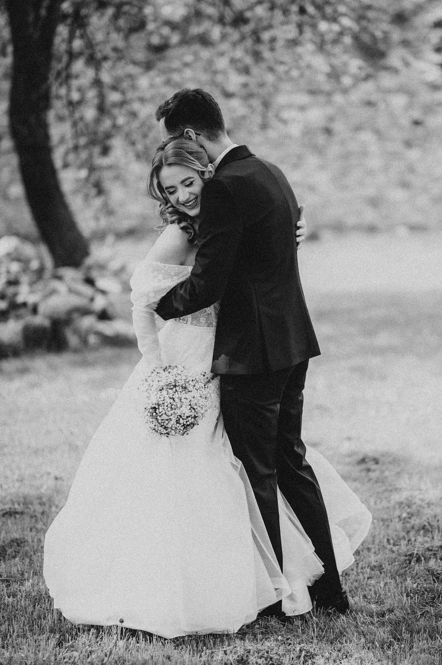 Black and white photo of a smiling bride in a white gown holding a bouquet, embraced by a groom in a dark suit outdoors.