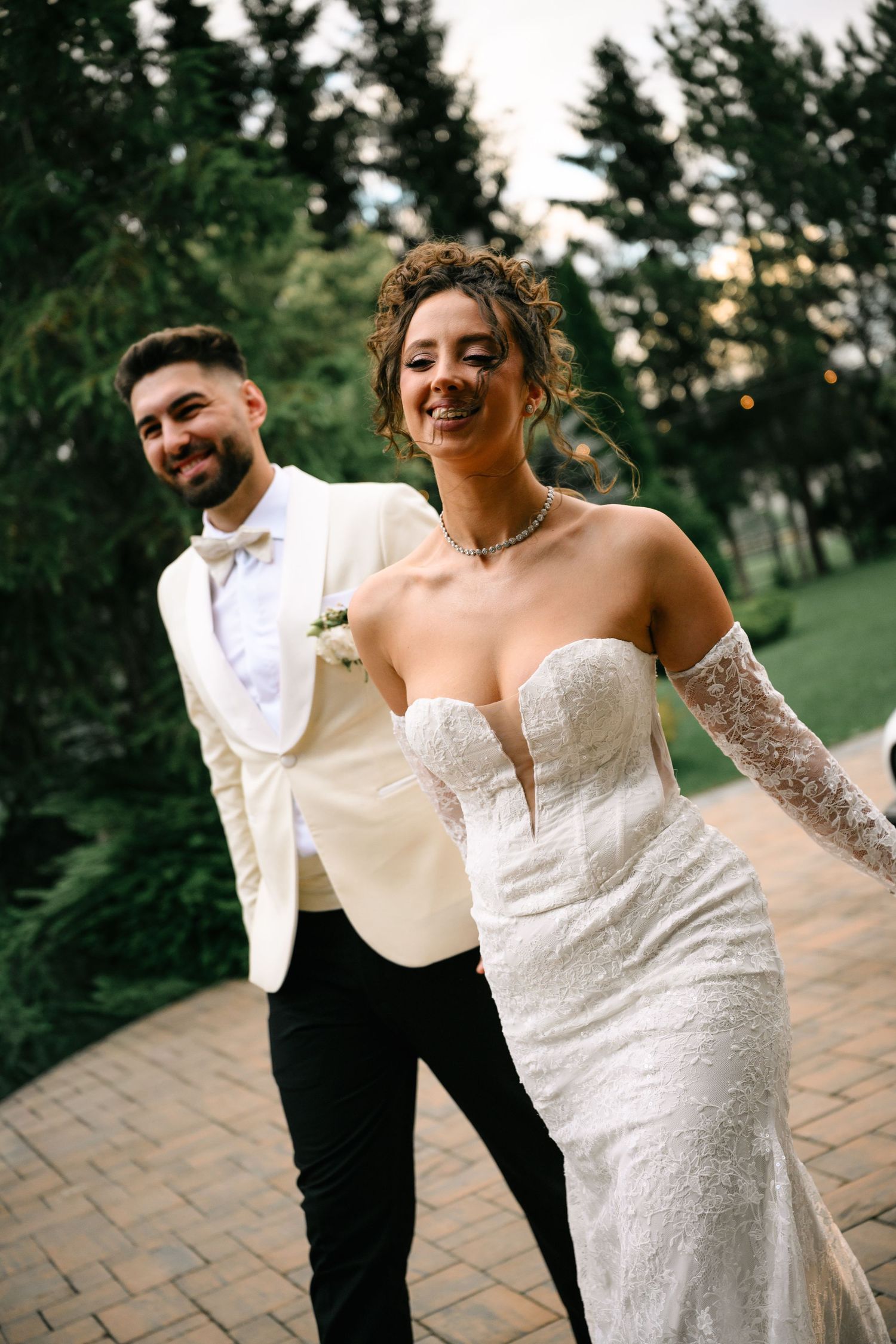 Smiling bride in a strapless lace gown with gloves leads groom in cream tuxedo along a garden pathway.