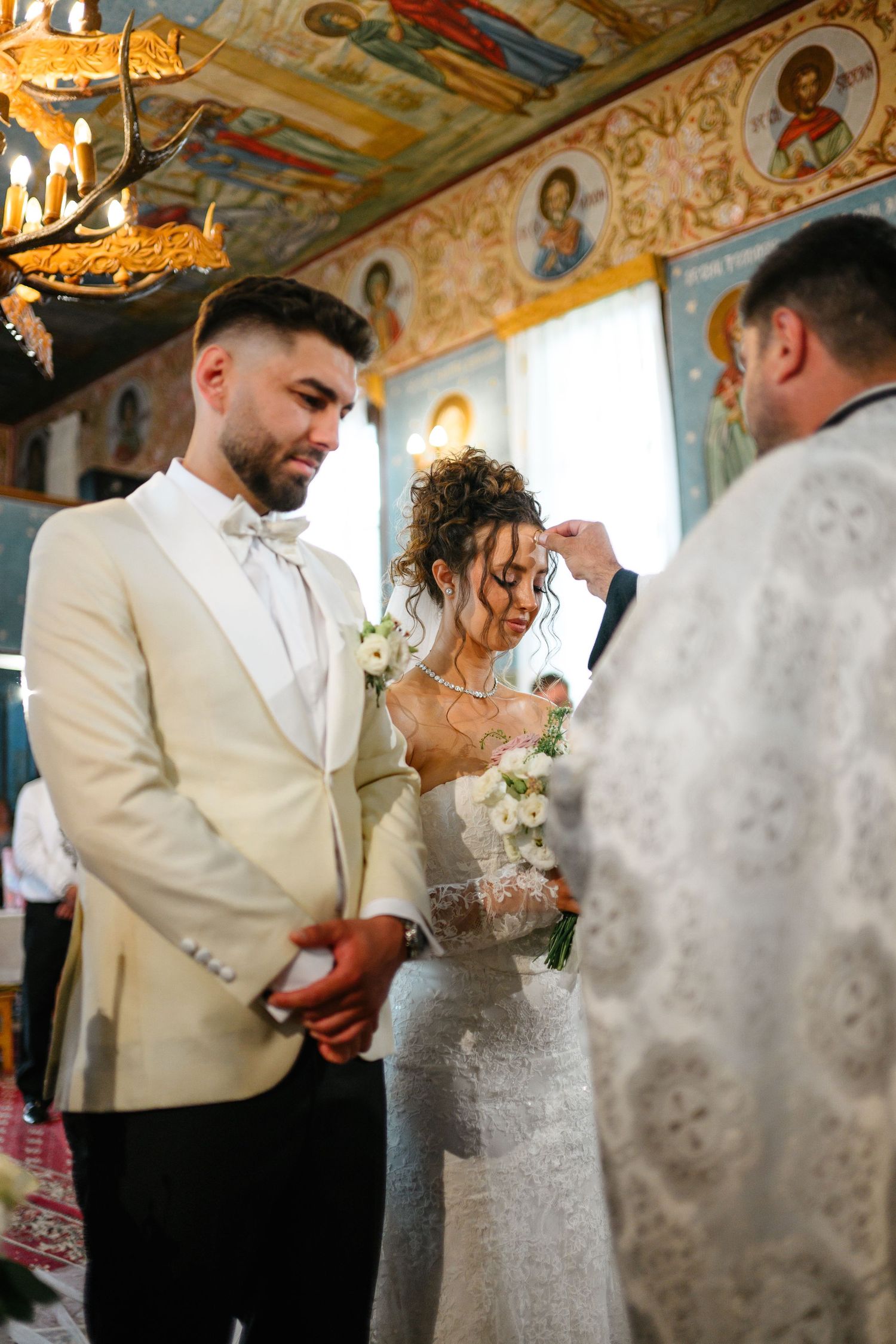 Bride and groom holding hands during Orthodox church wedding ceremony with ornate religious icons on walls.
