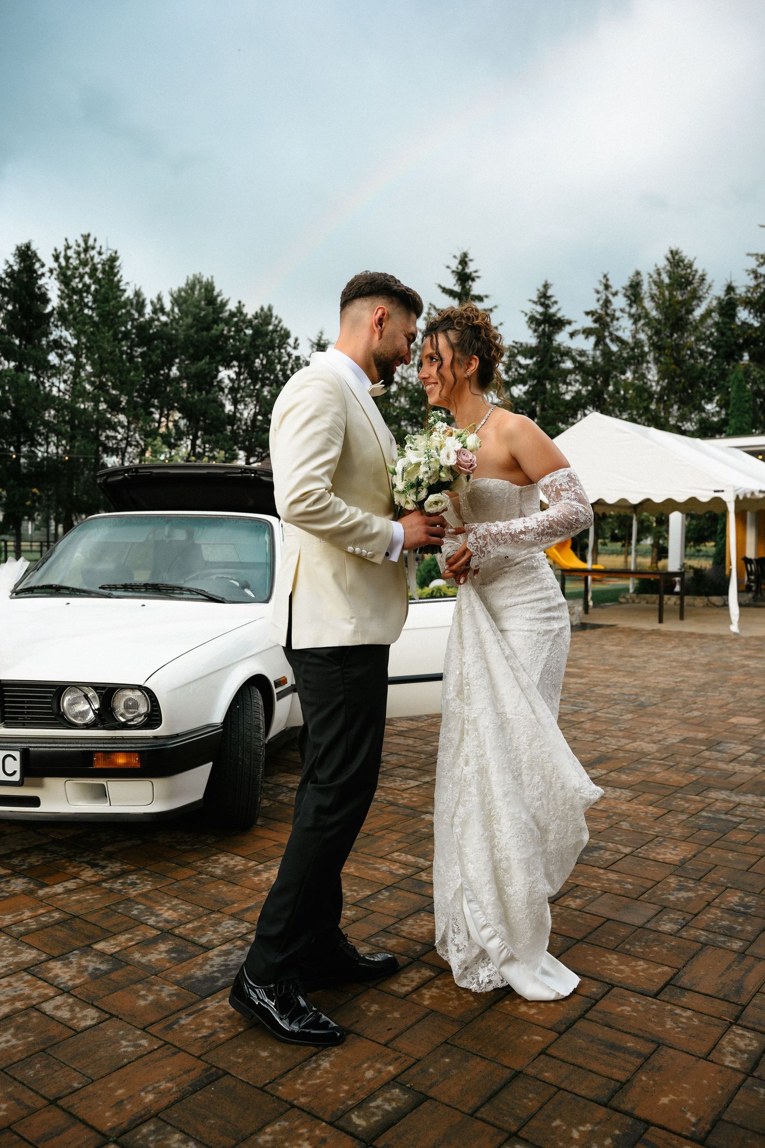 Bride and groom share a romantic moment beside a white vintage BMW with a rainbow in the cloudy sky backdrop.